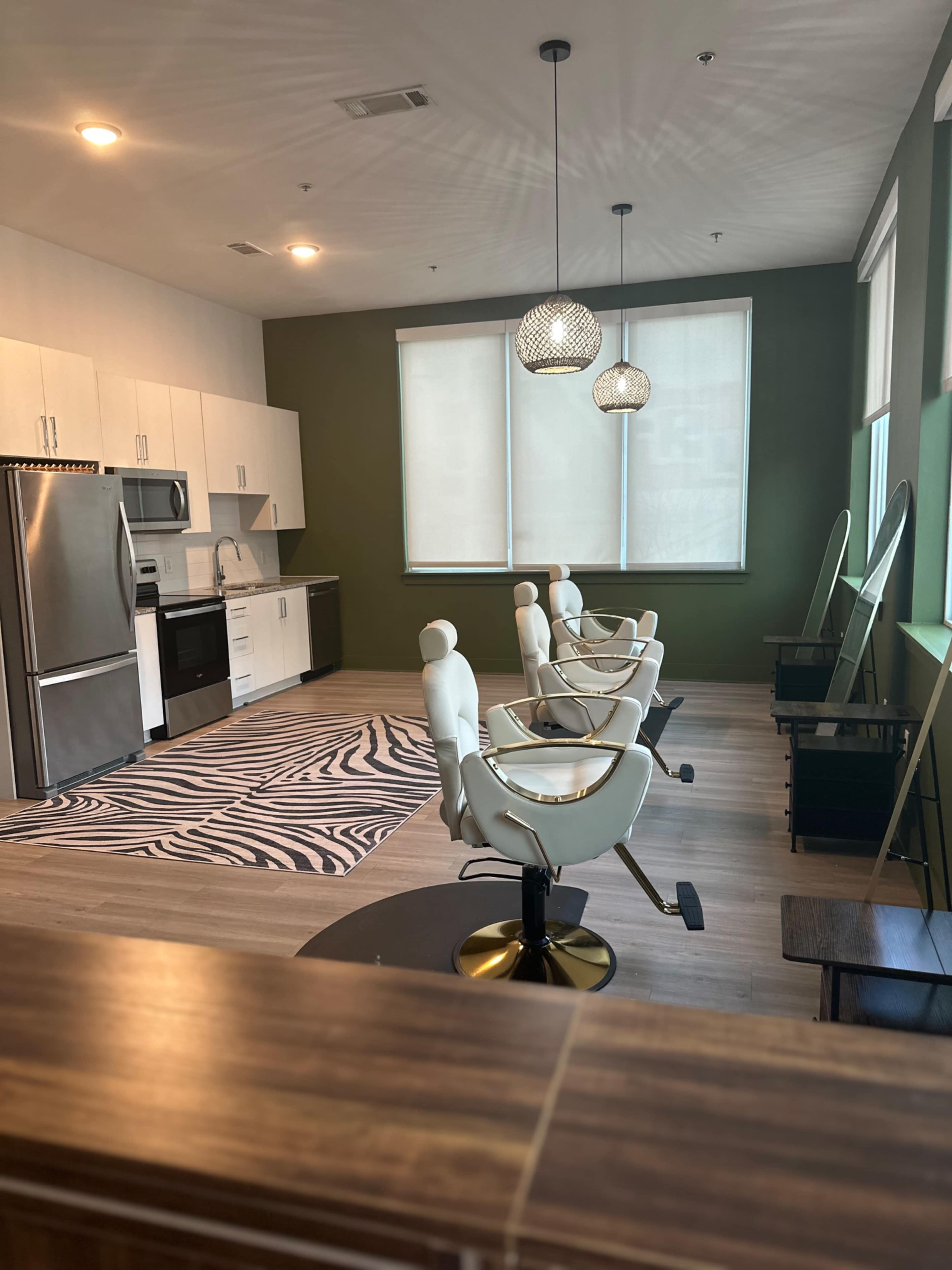 The image shows a modern salon interior with three styling chairs, a kitchen area, and large windows adorned with light-colored blinds.