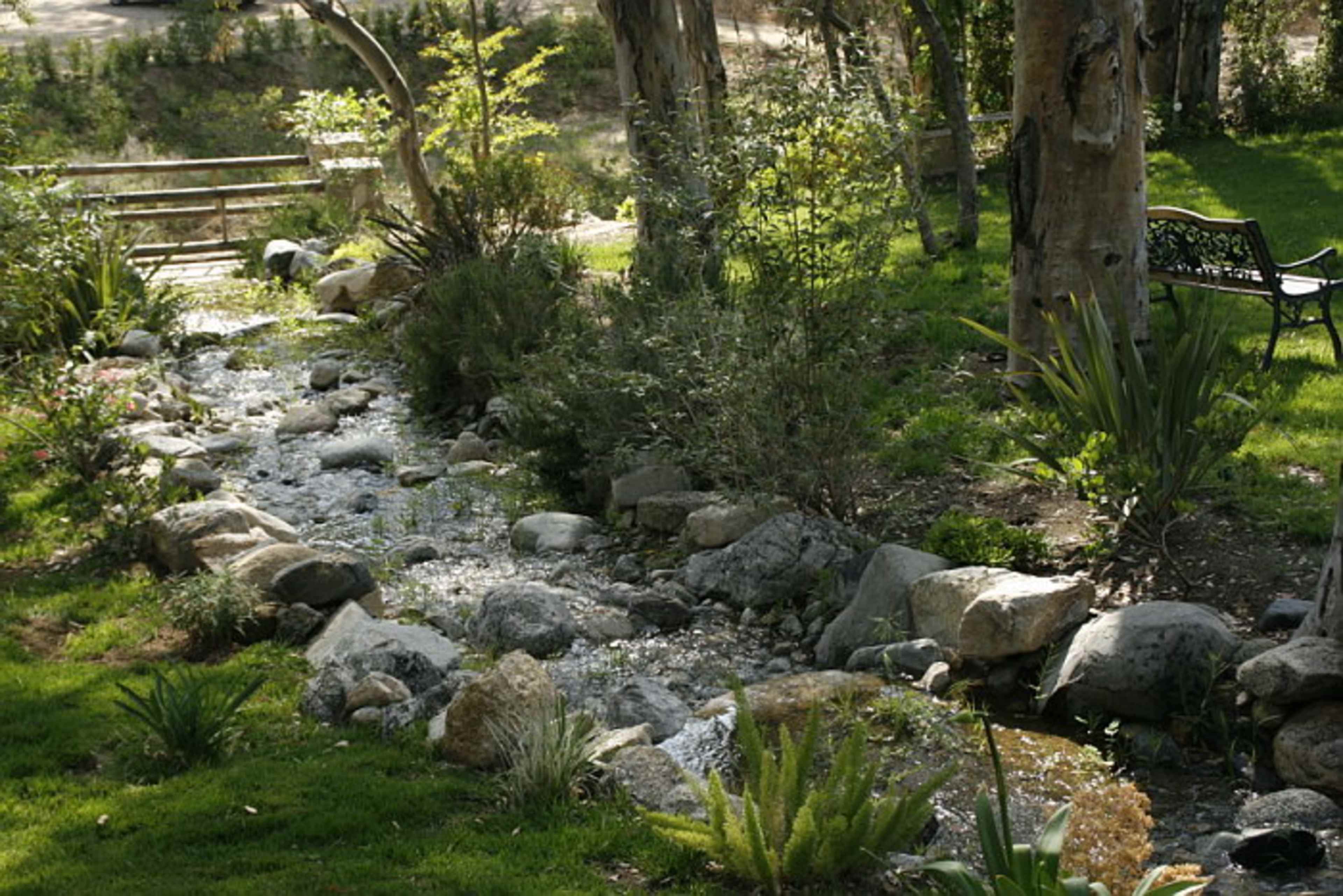 A narrow stream flows over rocks through a landscaped garden with greenery and a bench nearby.