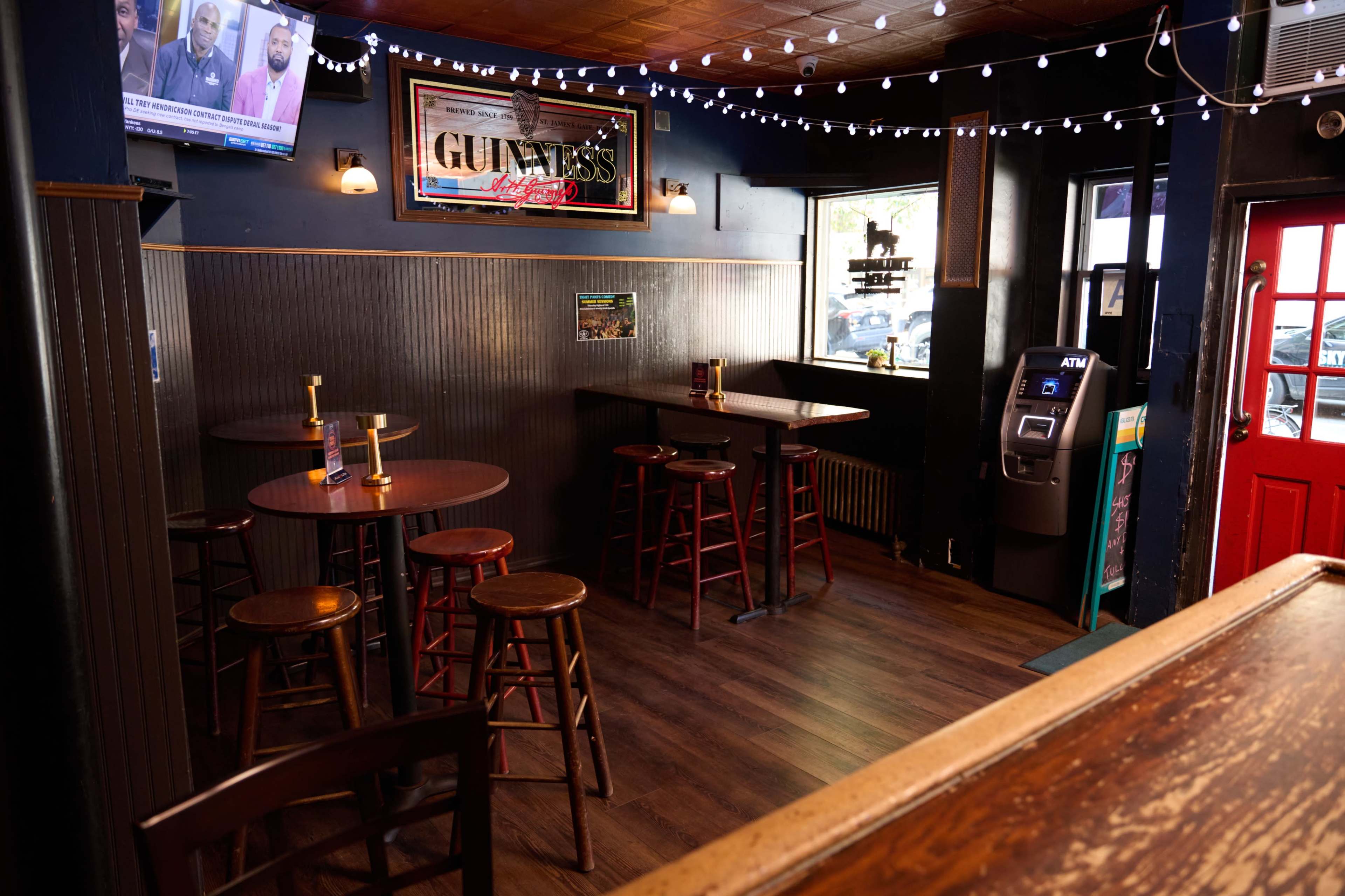 The interior of a pub features several wooden tables and chairs, a bar counter, and a brightly lit entrance with an ATM machine in the corner.