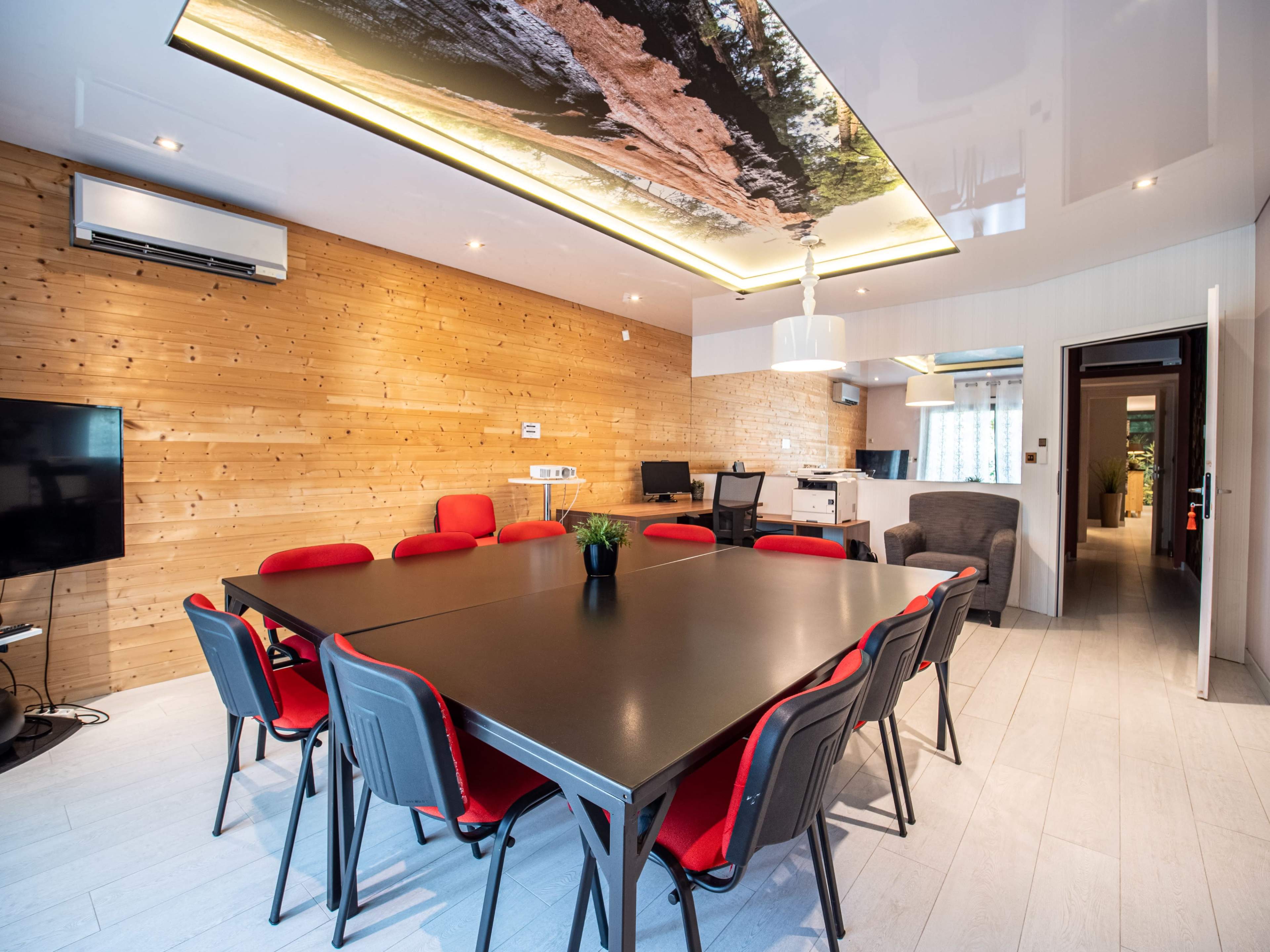 The image shows a modern meeting room with a large black table surrounded by red chairs, wooden walls, and a ceiling featuring a nature-themed design.