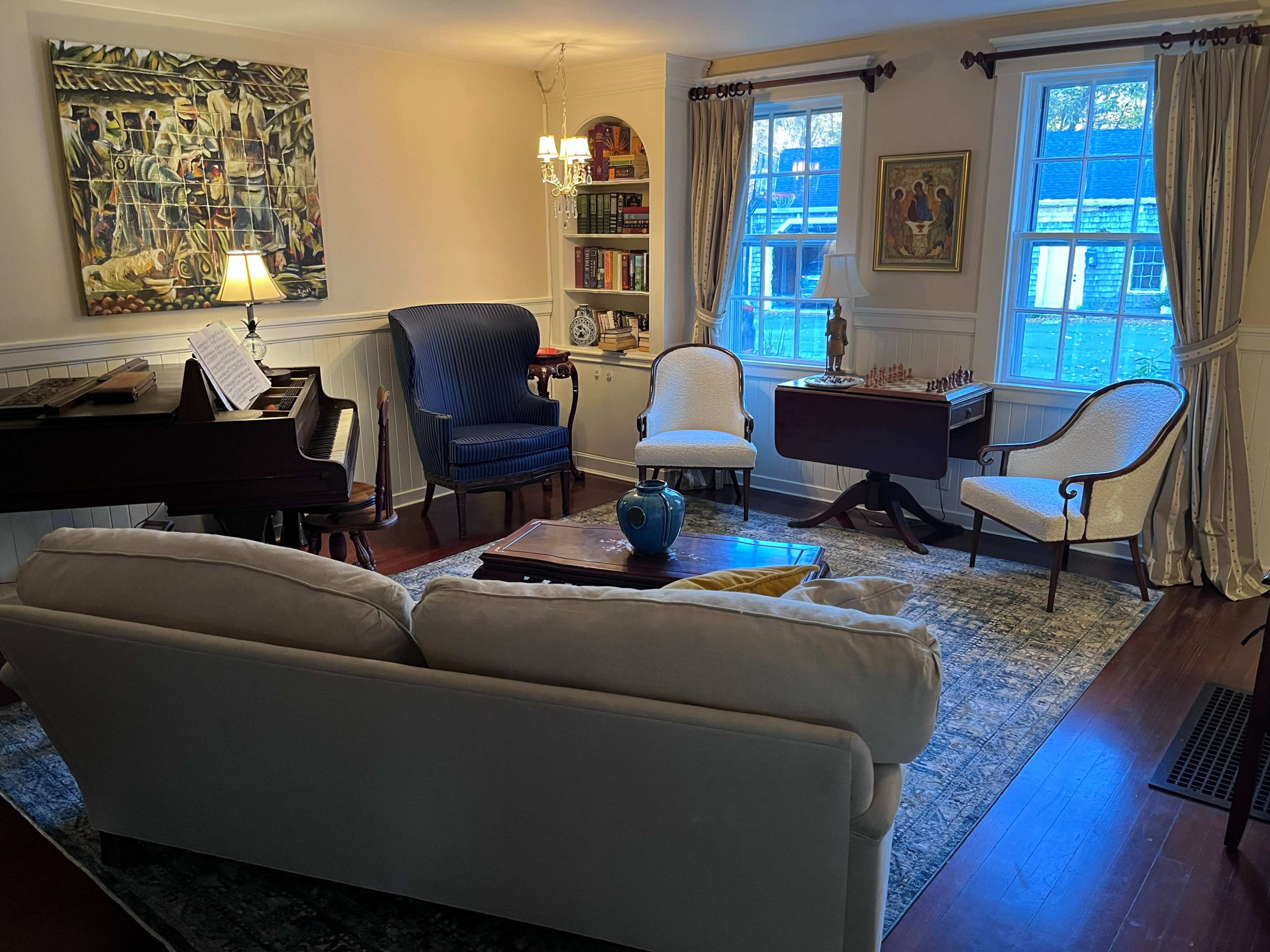 A living room features a piano, a chess table, two upholstered chairs, and a couch, with bookshelves and large windows in the background.