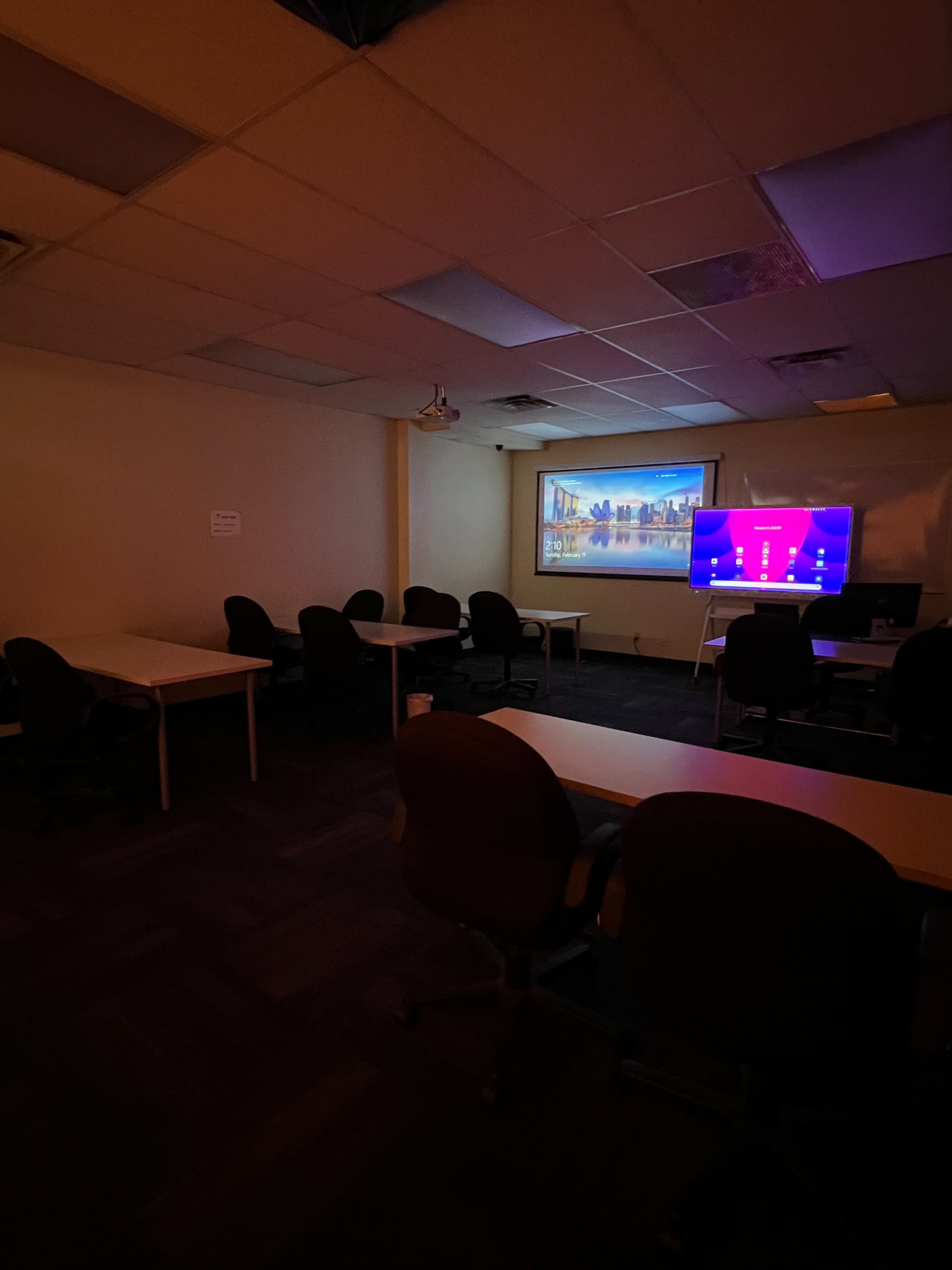 A dimly lit classroom features several empty tables and chairs, with a projector screen and a television displaying a digital interface on one wall.