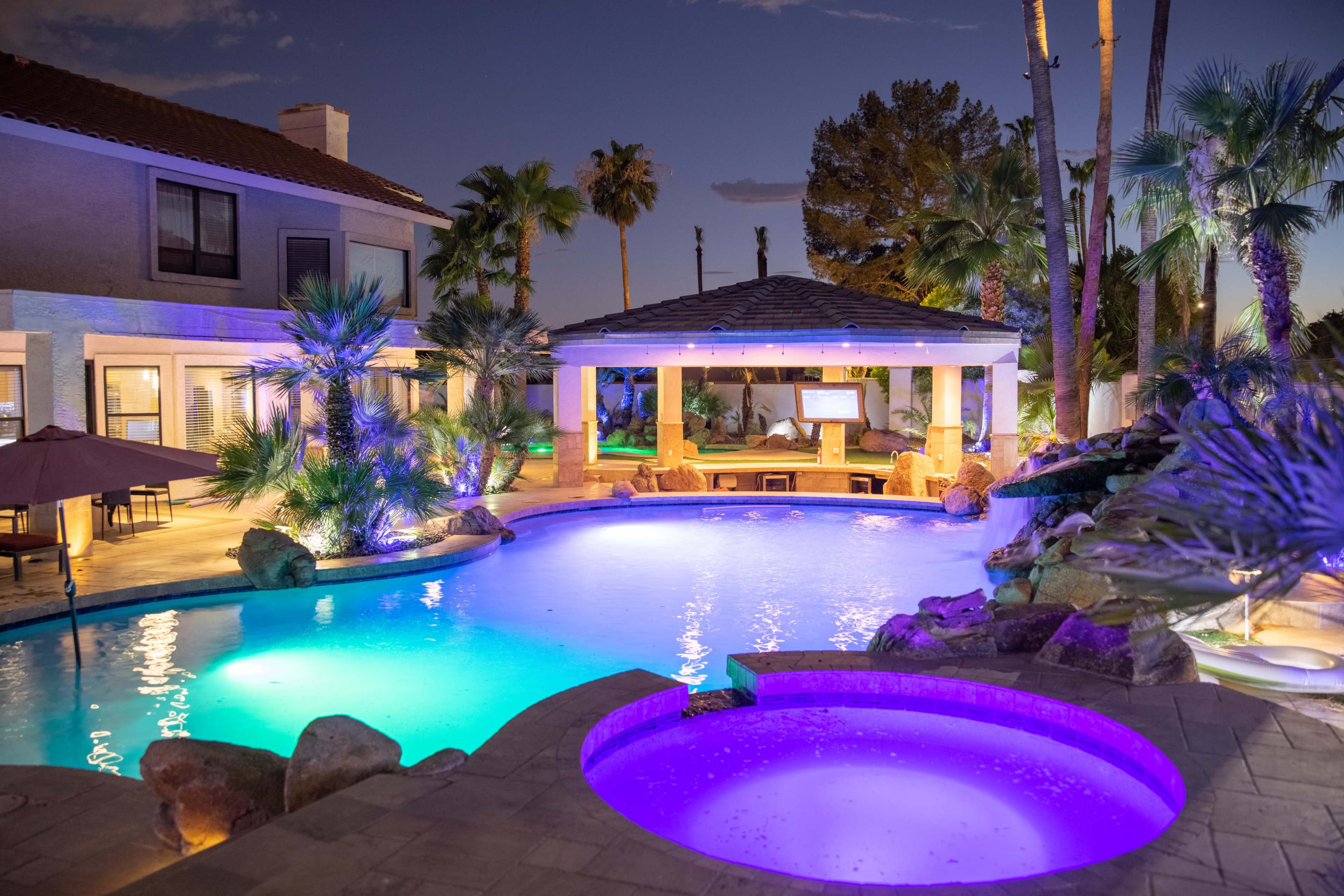 A luxurious backyard pool area with palm trees, a spa, and a covered lounge space, illuminated by colorful lights during twilight.