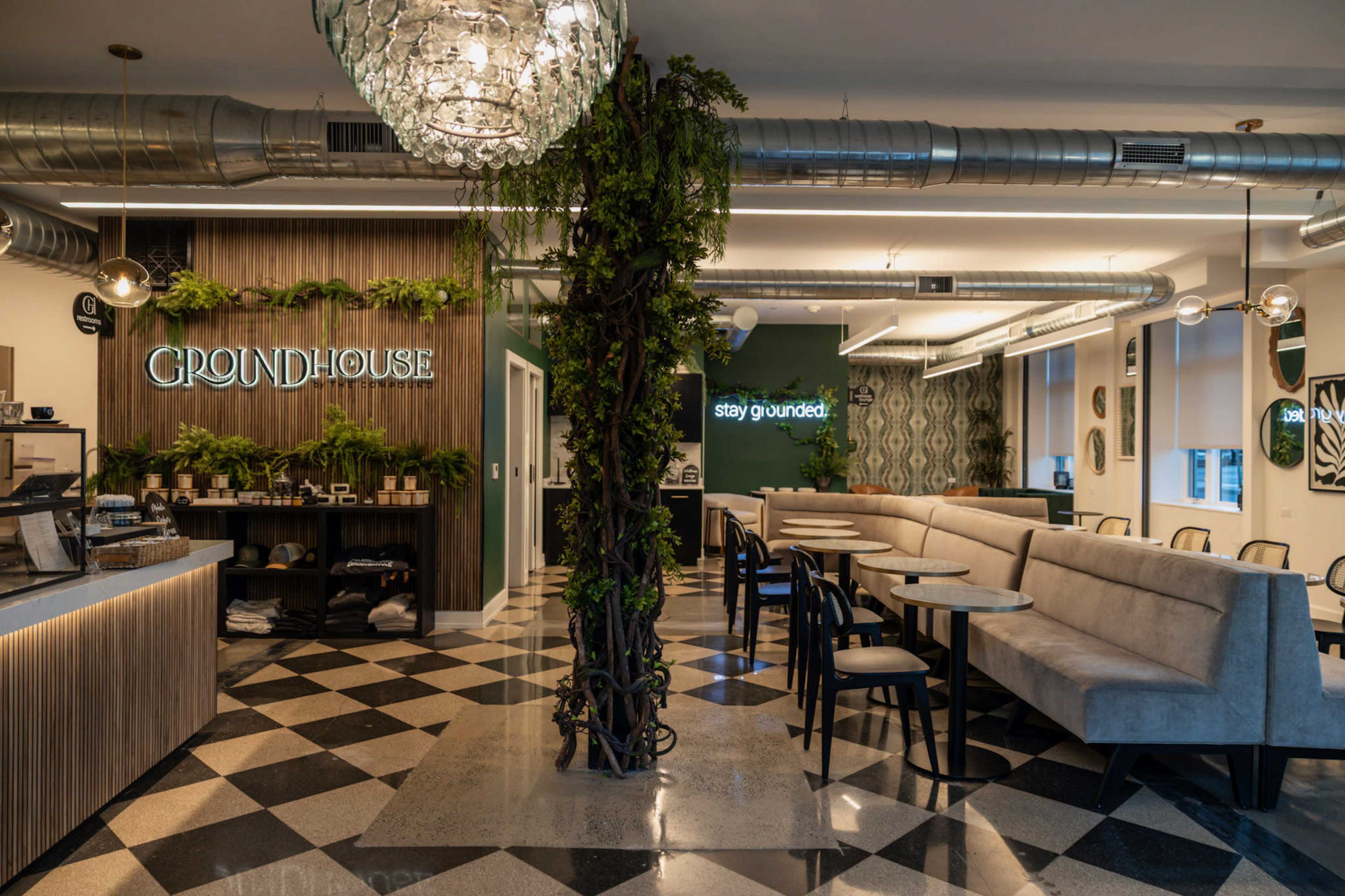 The image shows a modern café interior featuring a service counter on the left, plush seating areas, and decorative green plants along the walls and a central column.