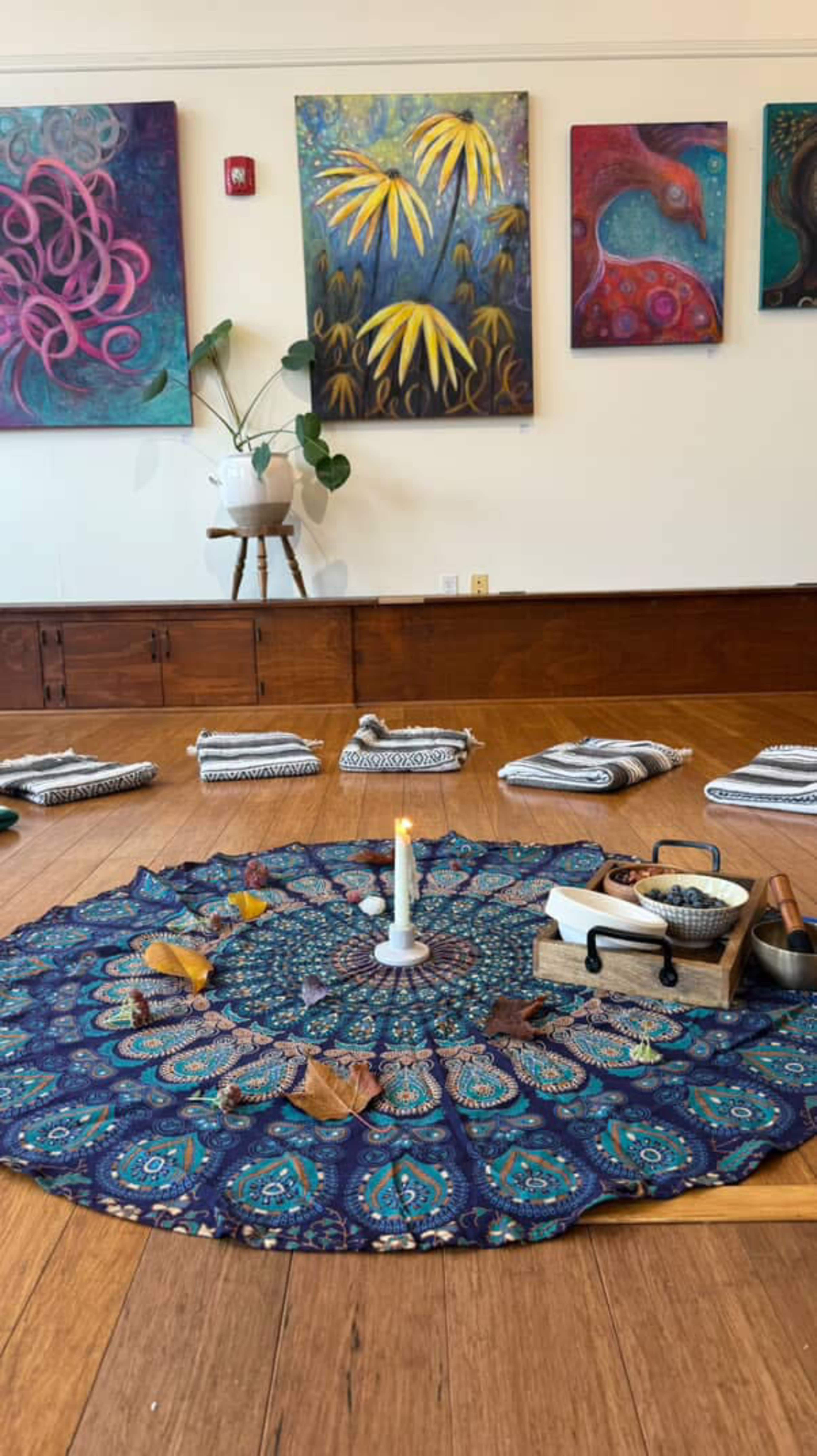A circular patterned mat is placed on the wooden floor, surrounded by neatly arranged blankets and a central candle, beneath colorful artistic paintings on the wall.