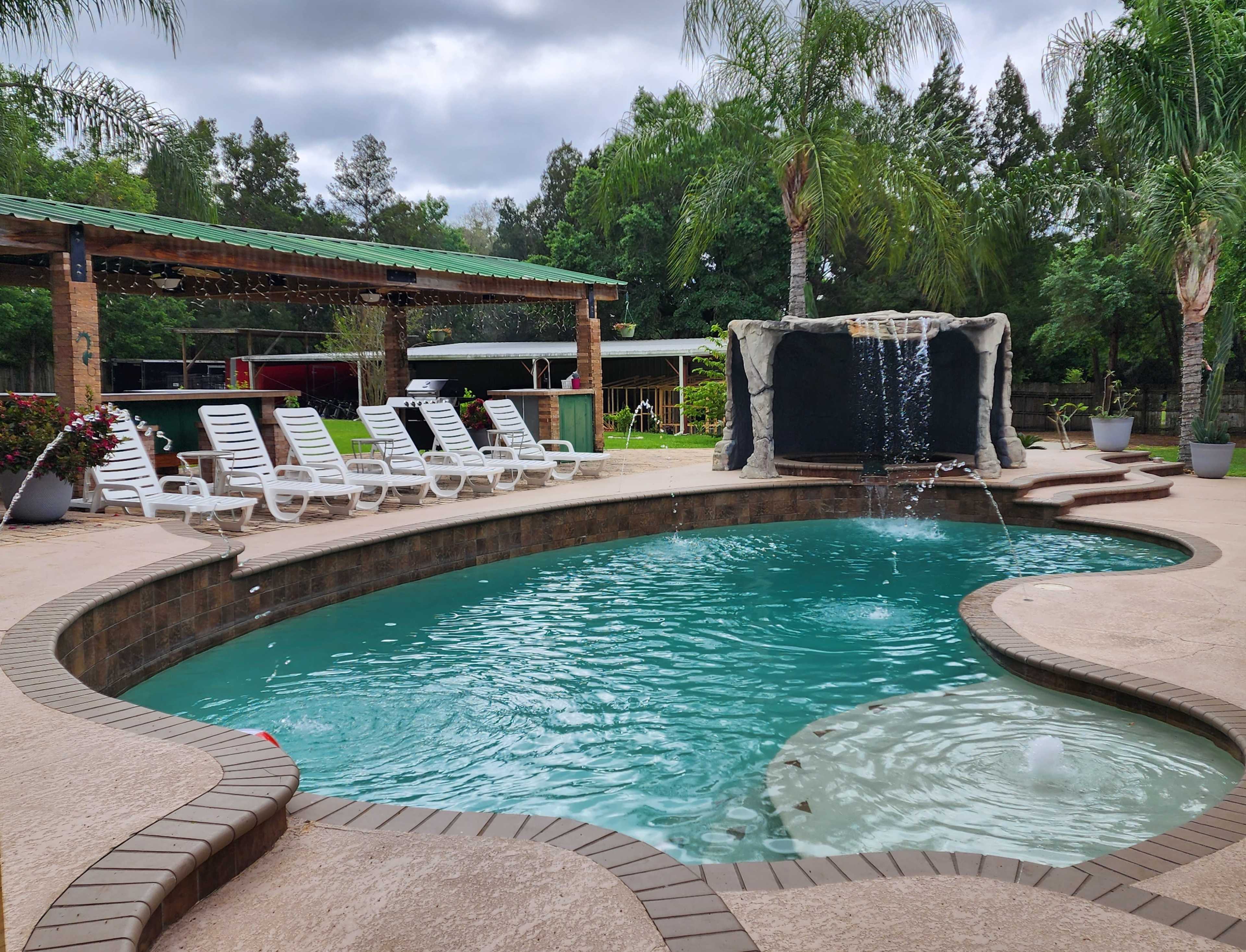 The image shows a swimming pool surrounded by lounge chairs, with a waterfall feature and palm trees in the background.