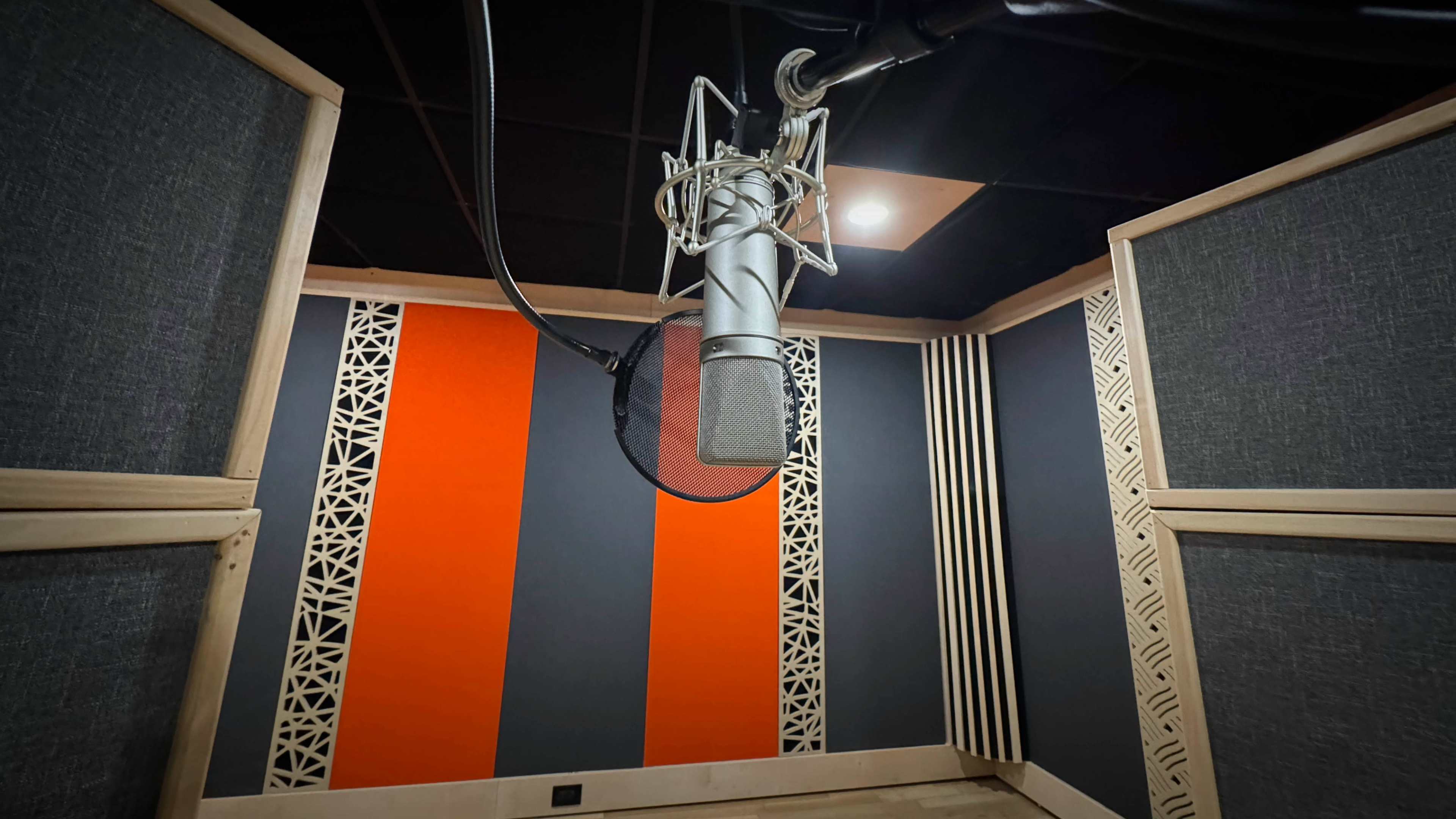 The image shows a microphone mounted on a stand in a soundproof recording studio with patterned walls in orange and gray.