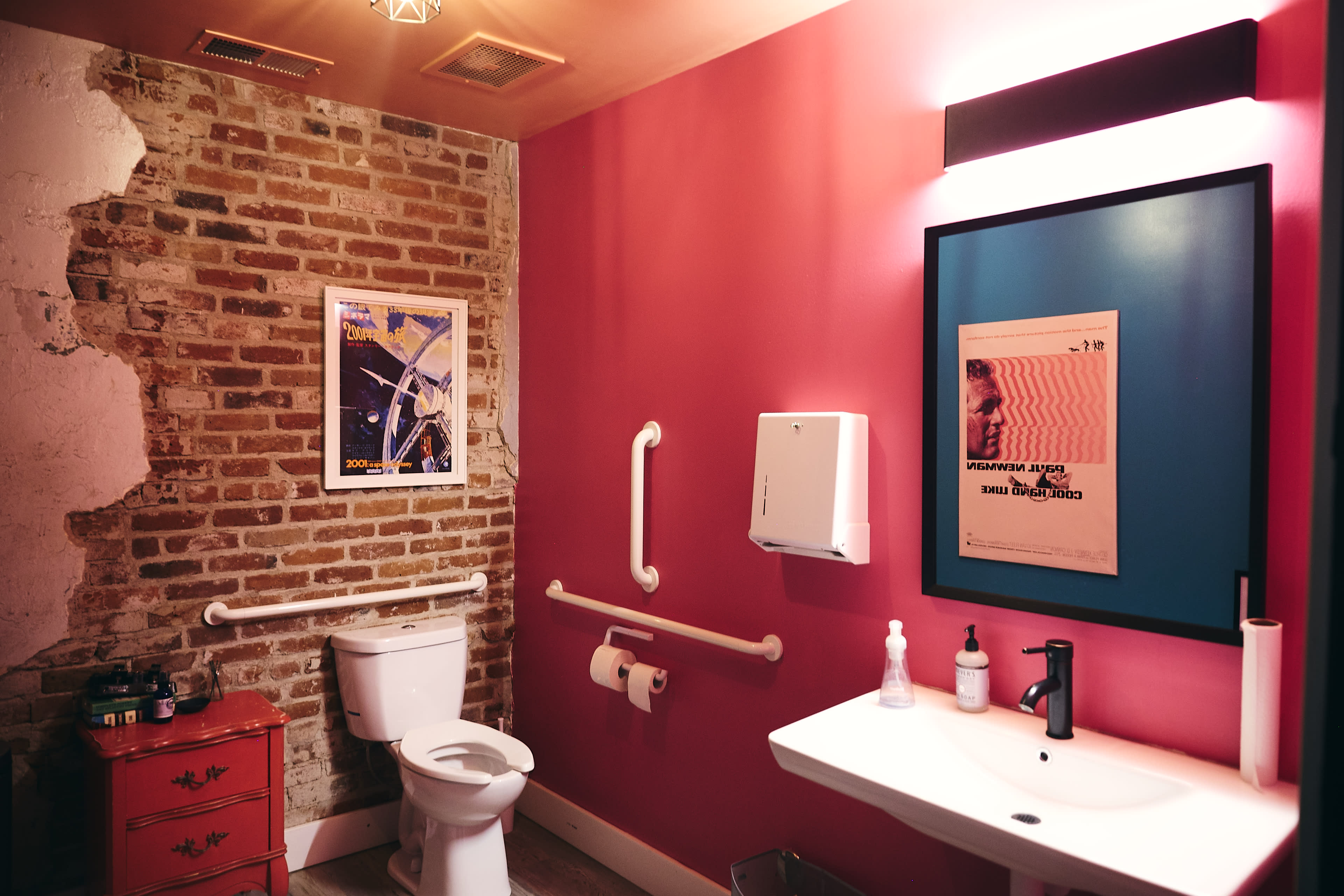 A modern bathroom featuring a toilet, a wall-mounted sink, a colorful poster, and a brick wall with exposed sections.