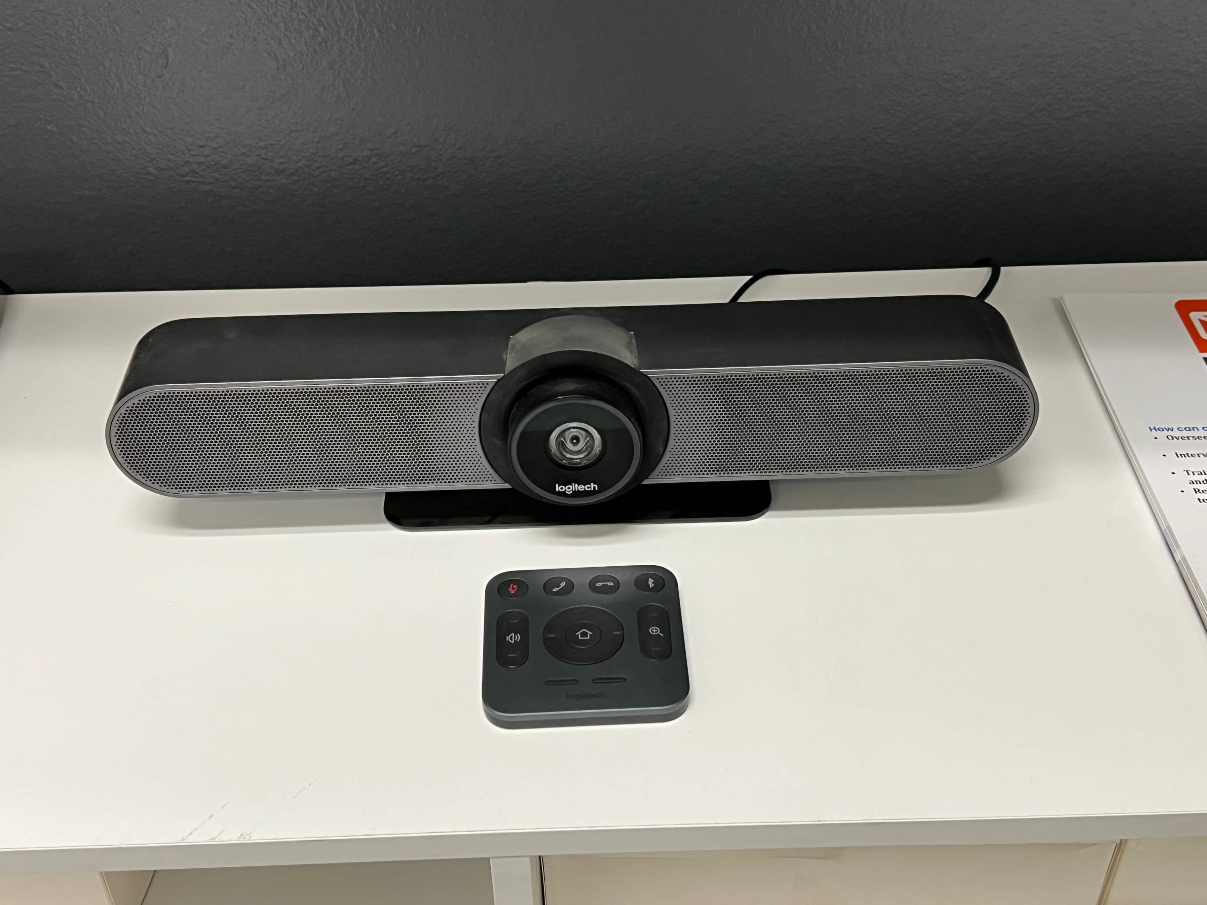 A Logitech speaker device with a circular camera lens sits on a white table next to a remote control.