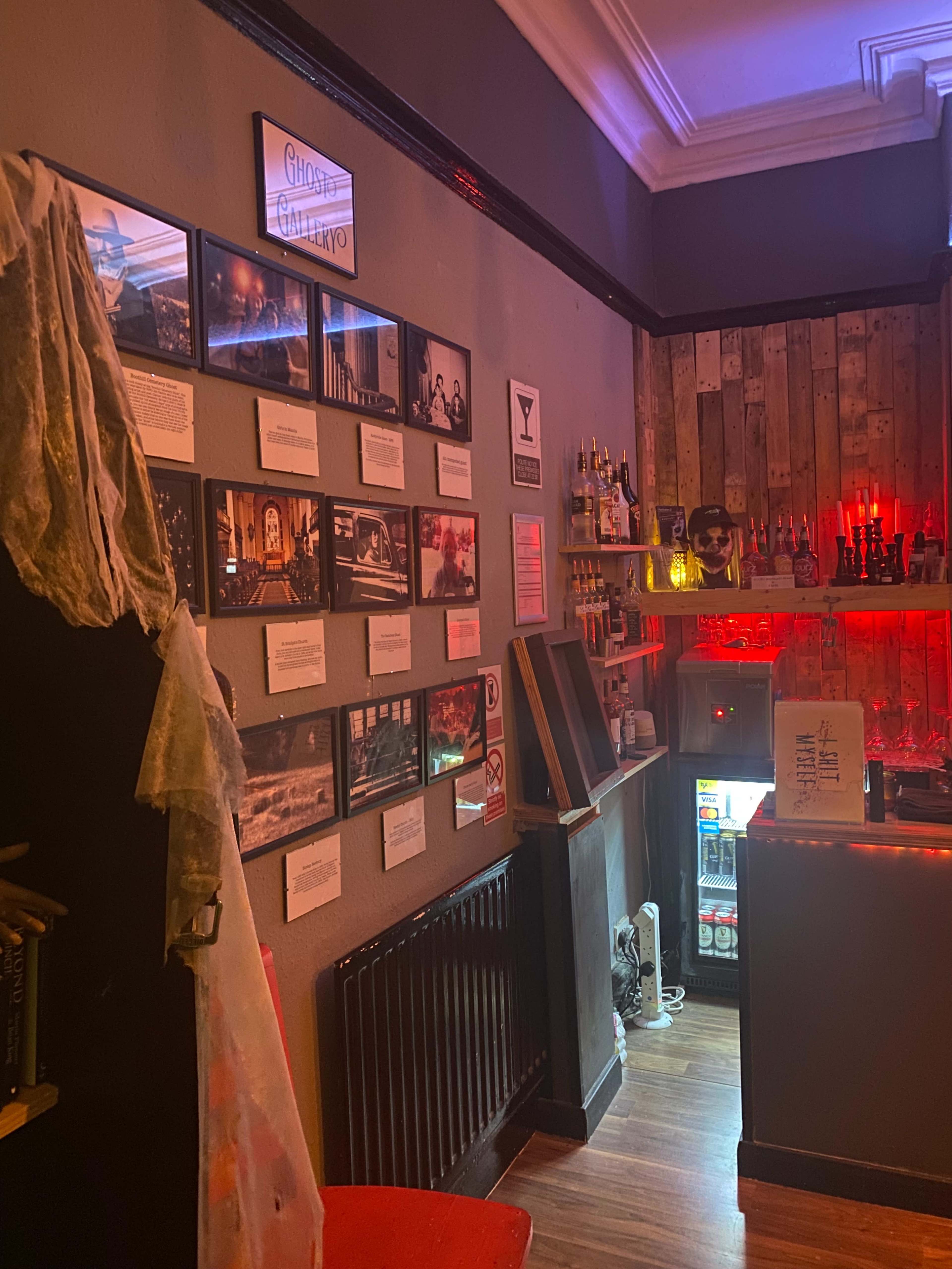The interior of a themed bar featuring a display of framed photographs on a wall, a wooden bar counter with an array of bottles, and a dimly lit atmosphere.
