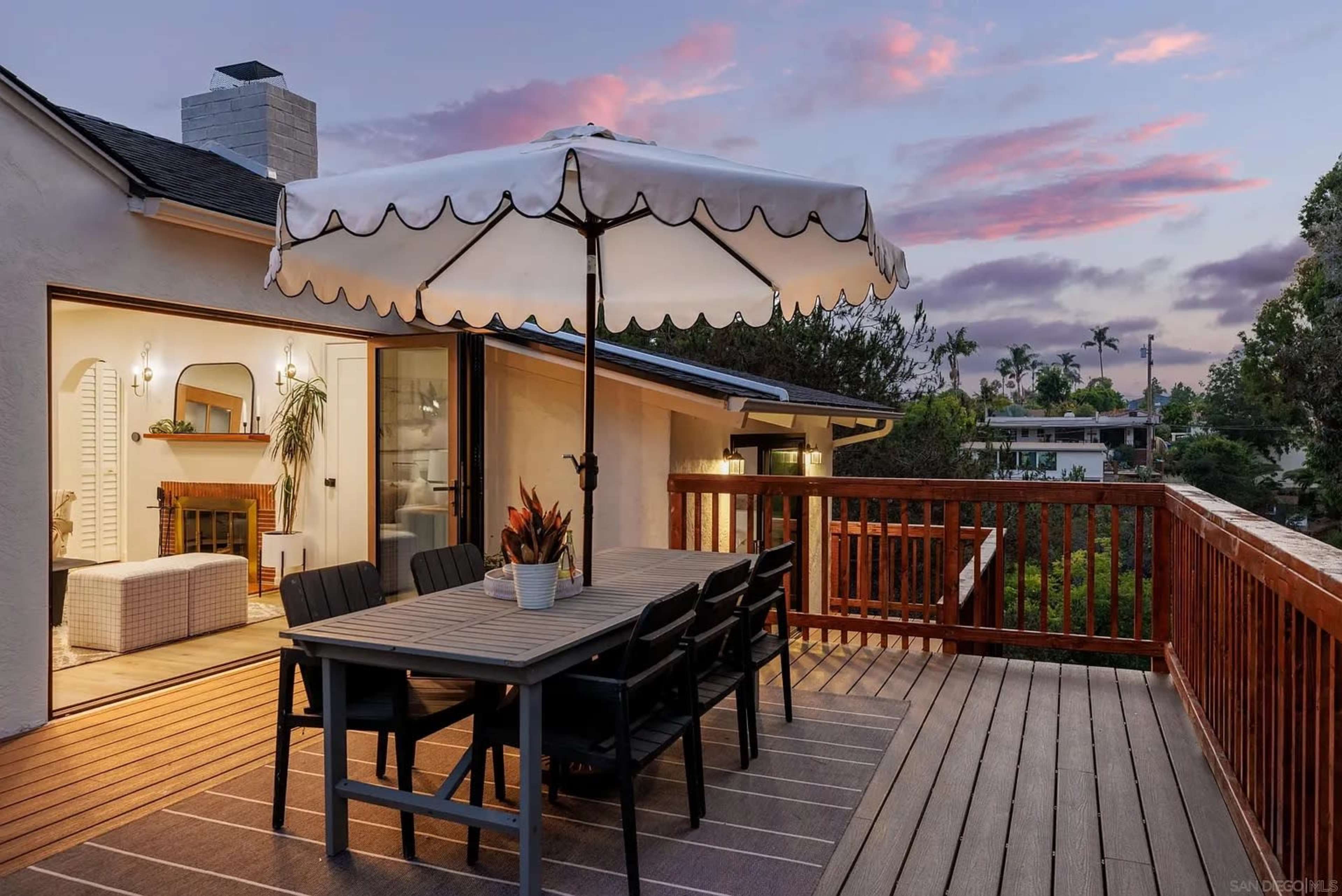 A wooden deck features a dining table with chairs under a large umbrella, overlooking a residential area at dusk.