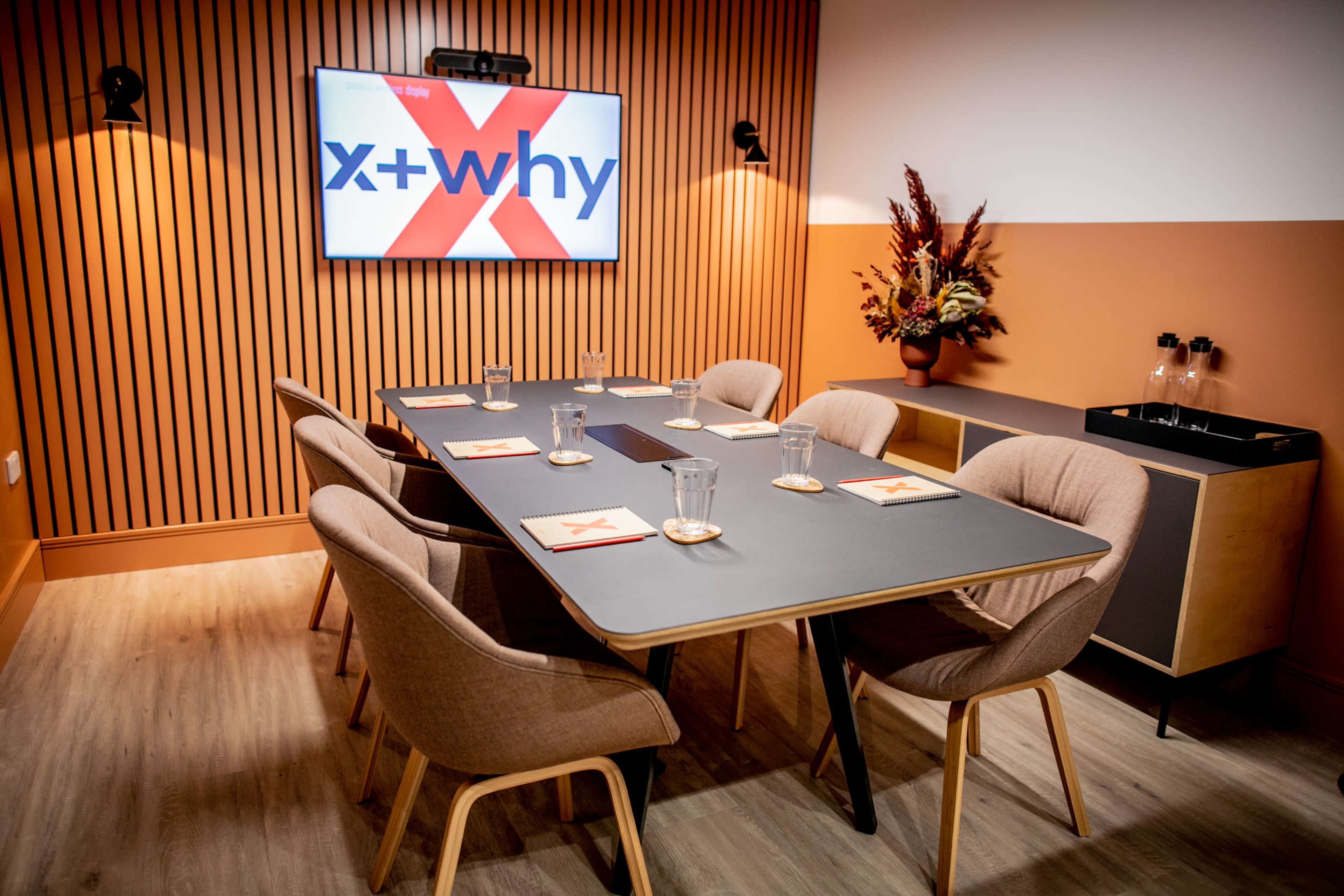 A modern conference room featuring a large table surrounded by six chairs, with a screen displaying "x+why" on the wall and decorative elements in the background.
