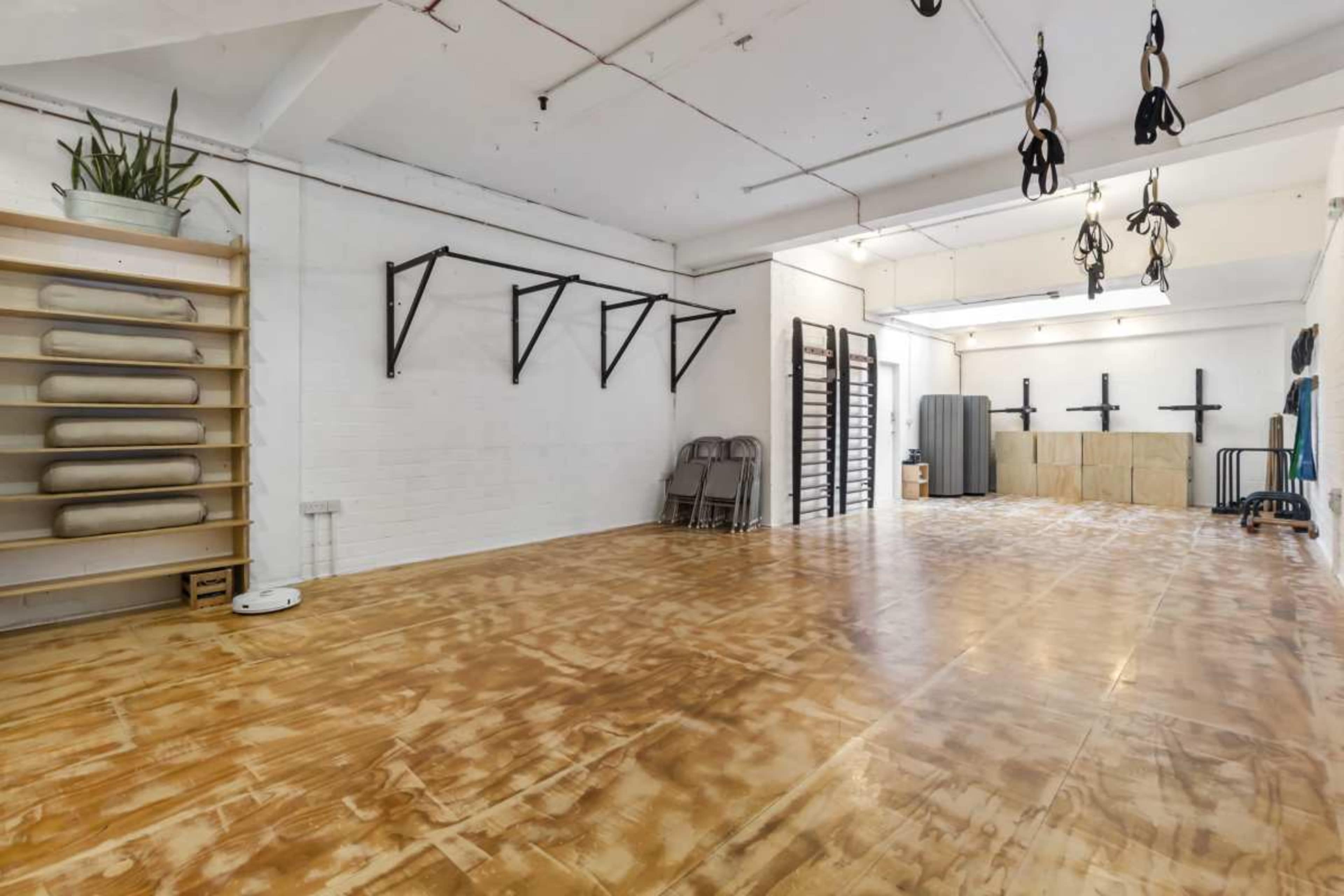 The image showcases a spacious, empty studio with wooden flooring, wall-mounted workout equipment, and neatly arranged mats on a shelf.