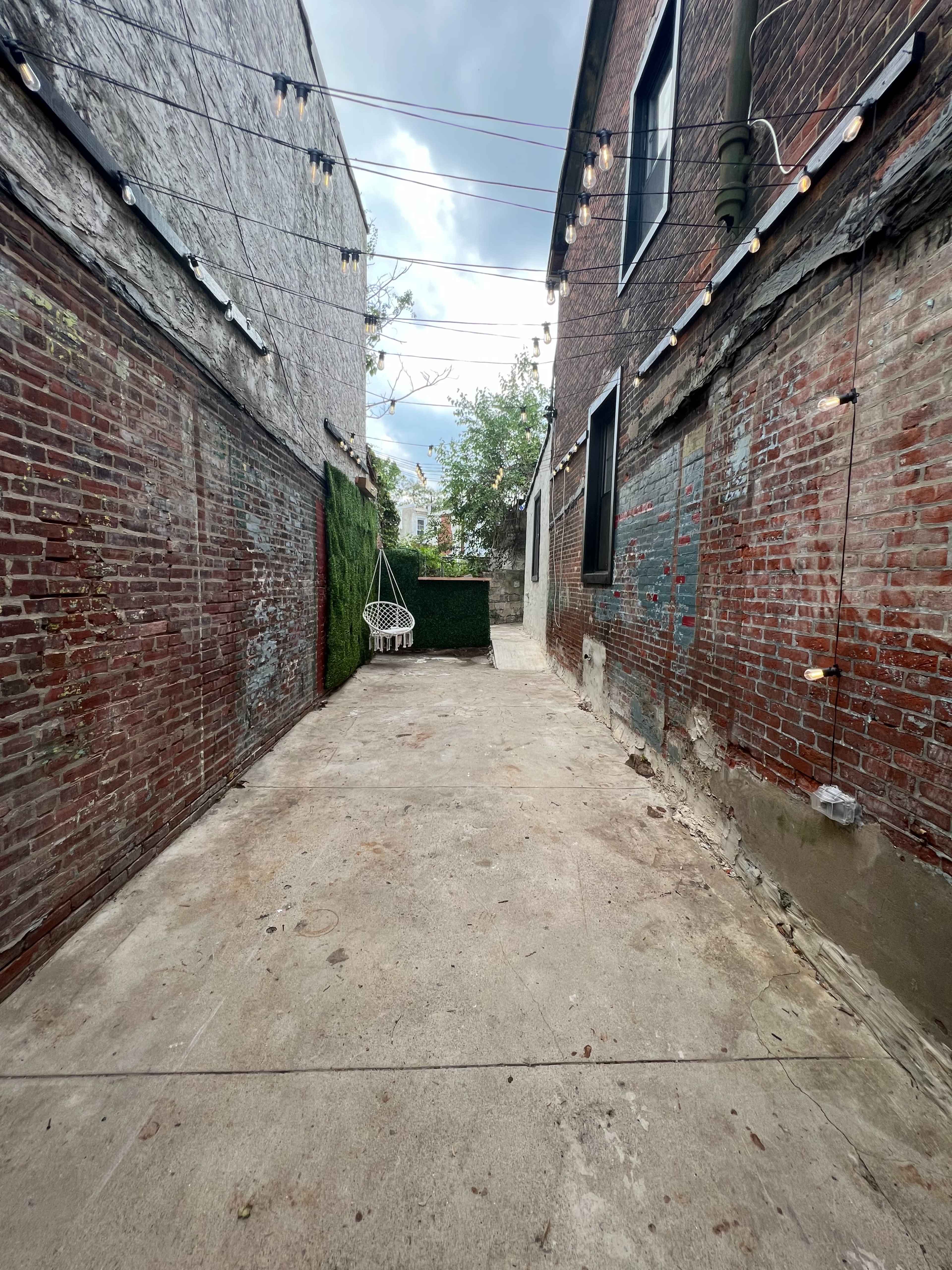 A narrow alleyway flanked by brick walls, leading to a concrete space with a hanging chair and greenery at the far end.