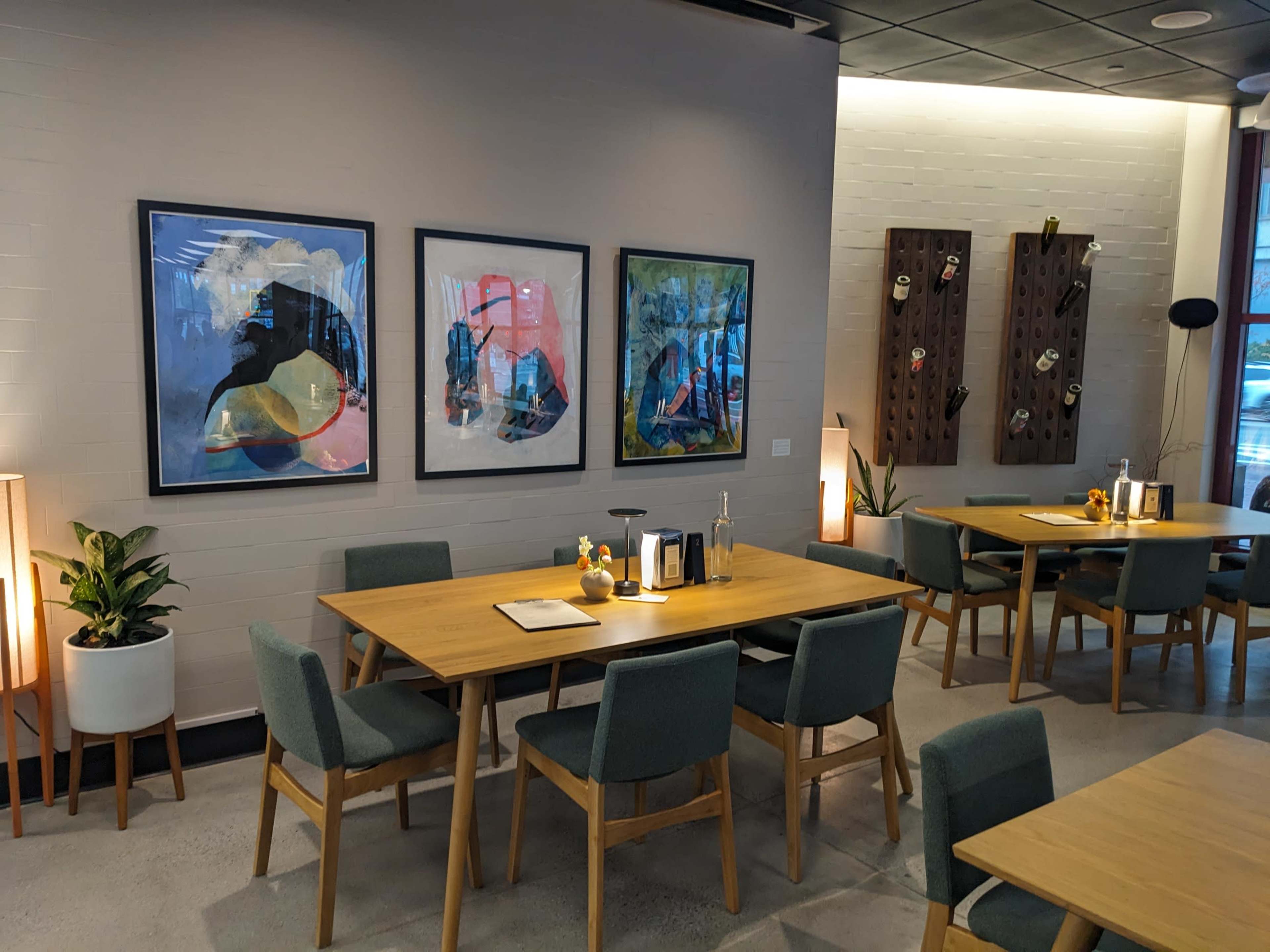 The image shows a modern dining area featuring wooden tables, green chairs, and framed artwork on the walls.