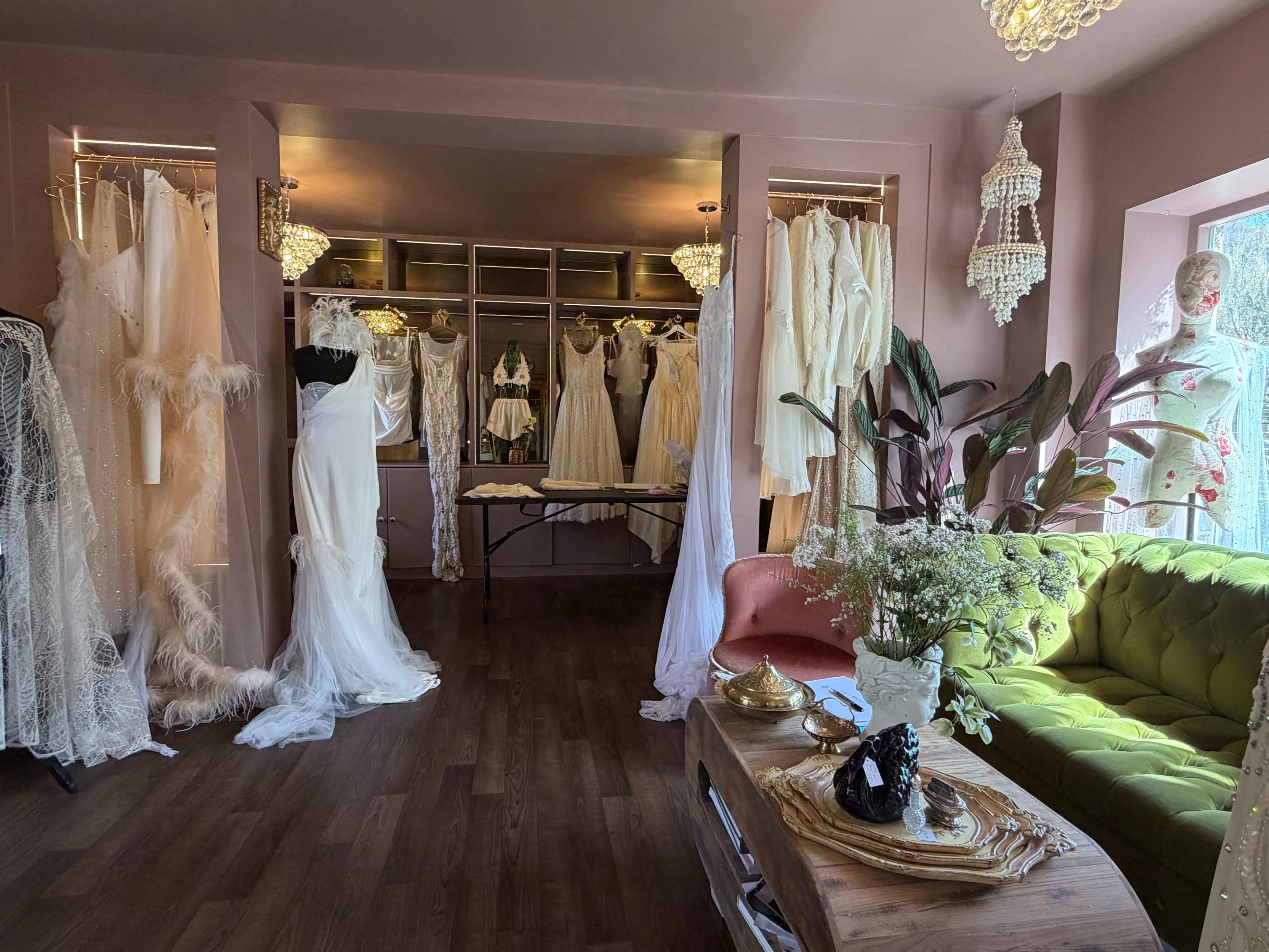 The image shows a boutique interior with various white dresses displayed on racks, a green sofa, and decorative chandeliers.
