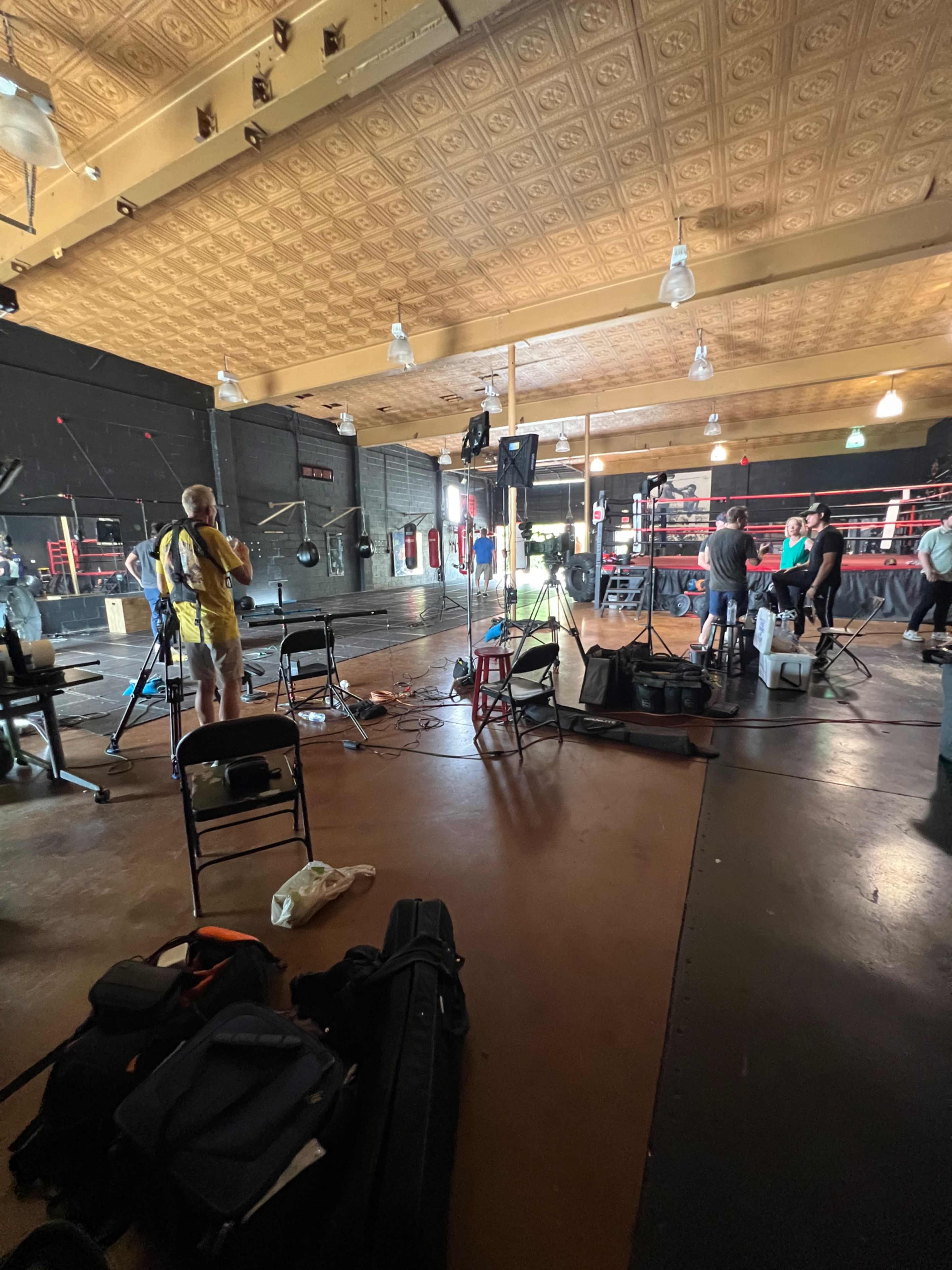 The scene shows a boxing gym set up for an event, with cameras, lighting equipment, and crew members preparing around the boxing ring.