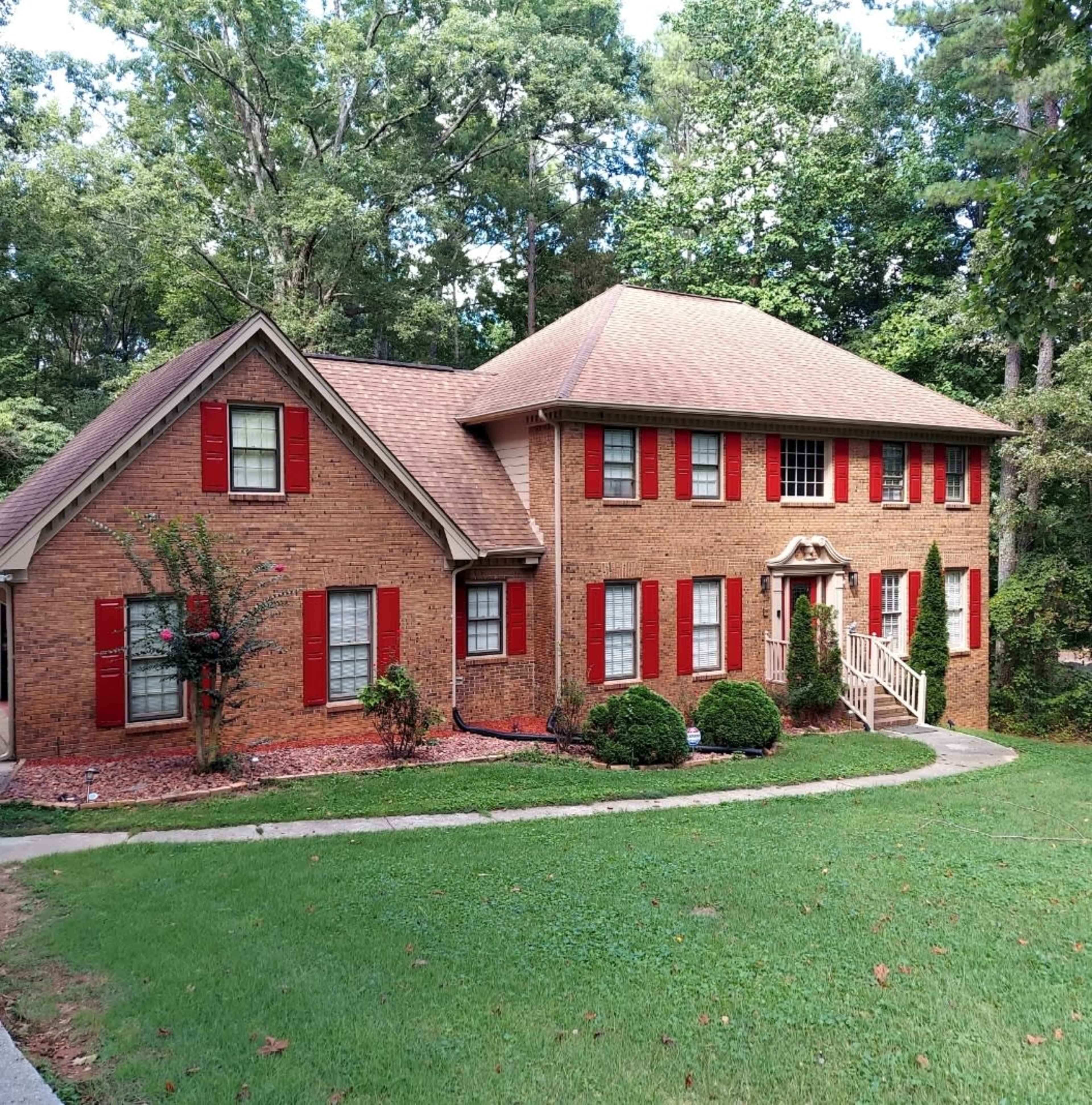 A two-story brick house with red shutters and a front porch is surrounded by a landscaped yard and trees.