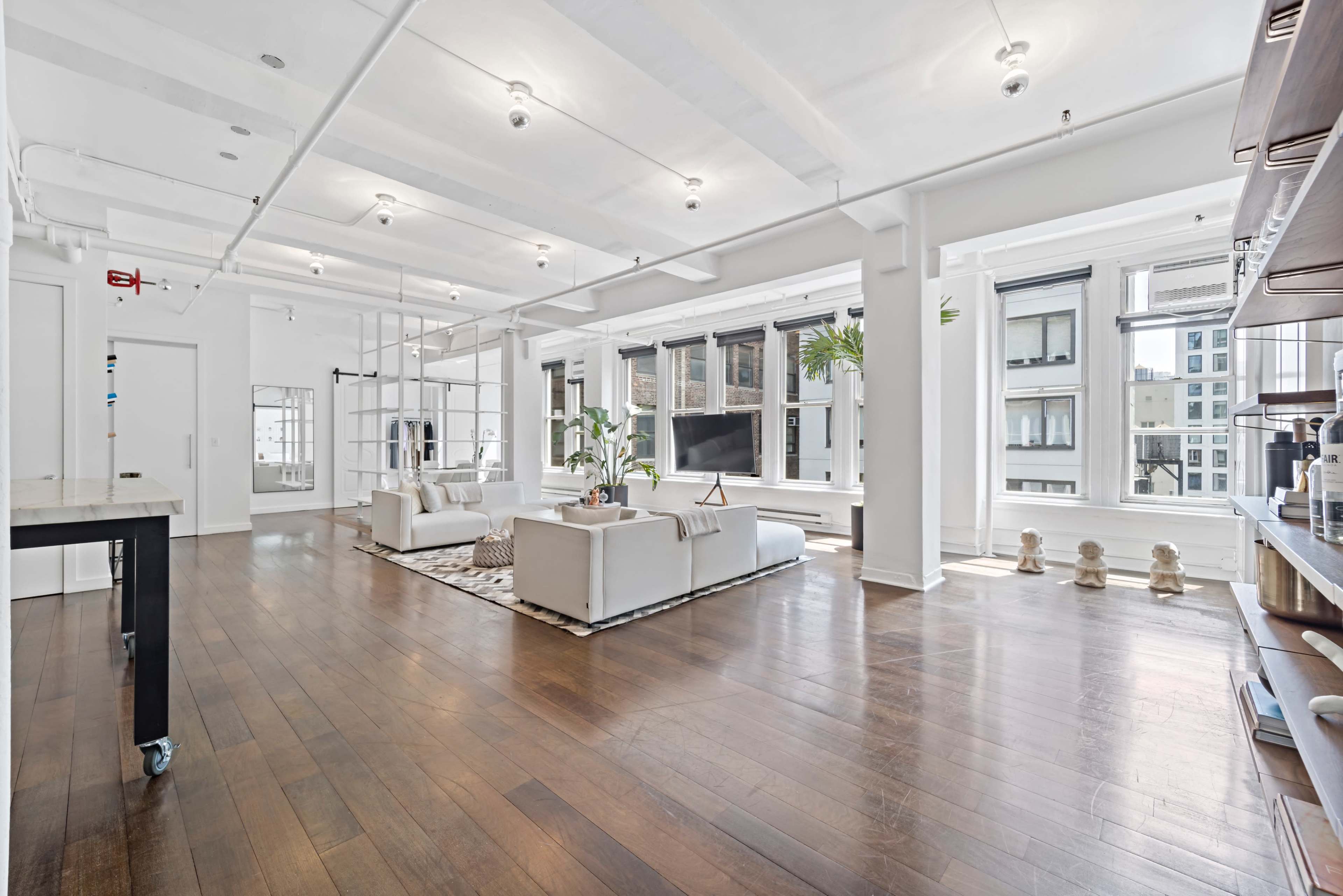 A spacious, brightly lit loft features white furnishings, large windows, and hardwood floors.