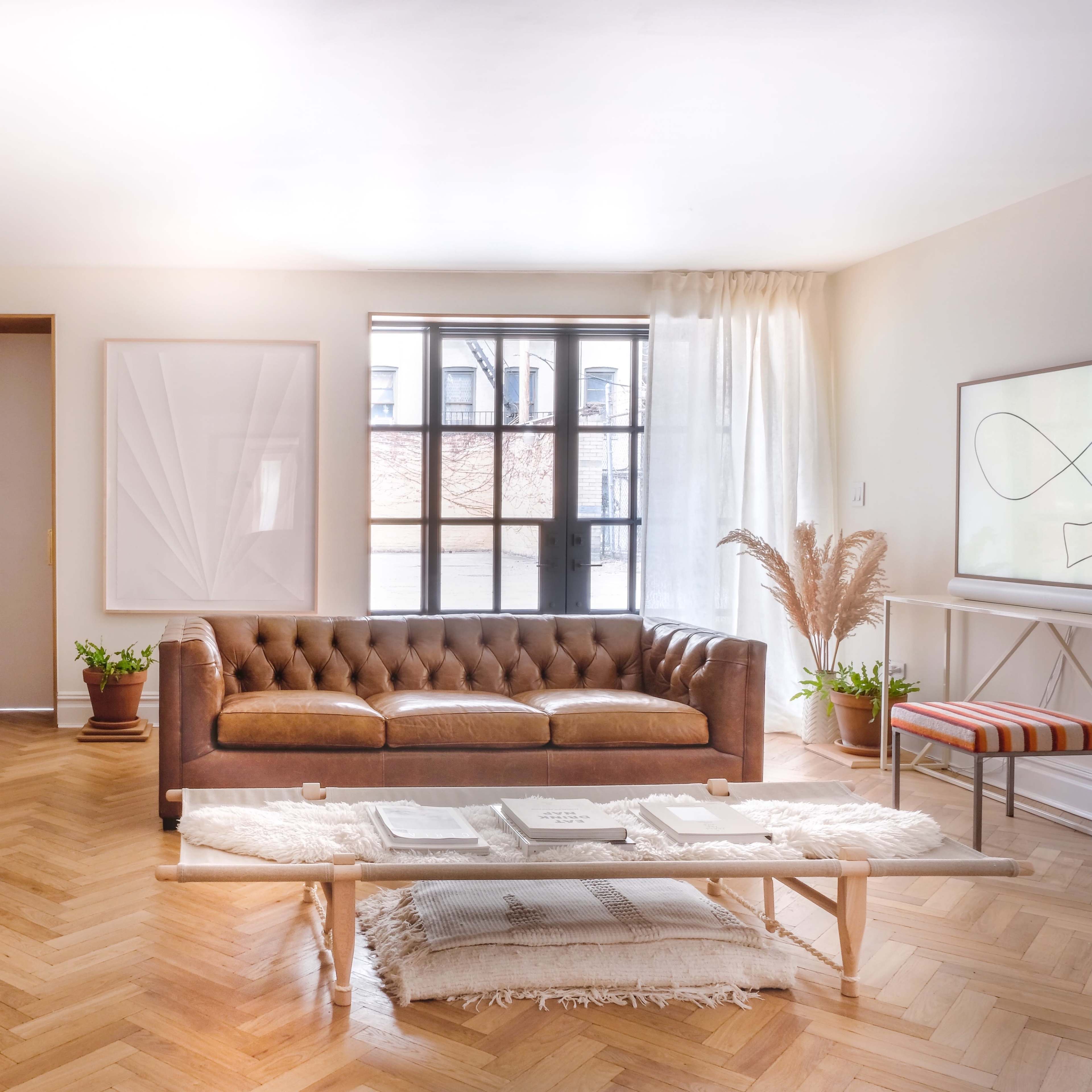 A modern living room features a brown leather tufted sofa, a wooden coffee table with decorative items, and a large window allowing natural light to fill the space.