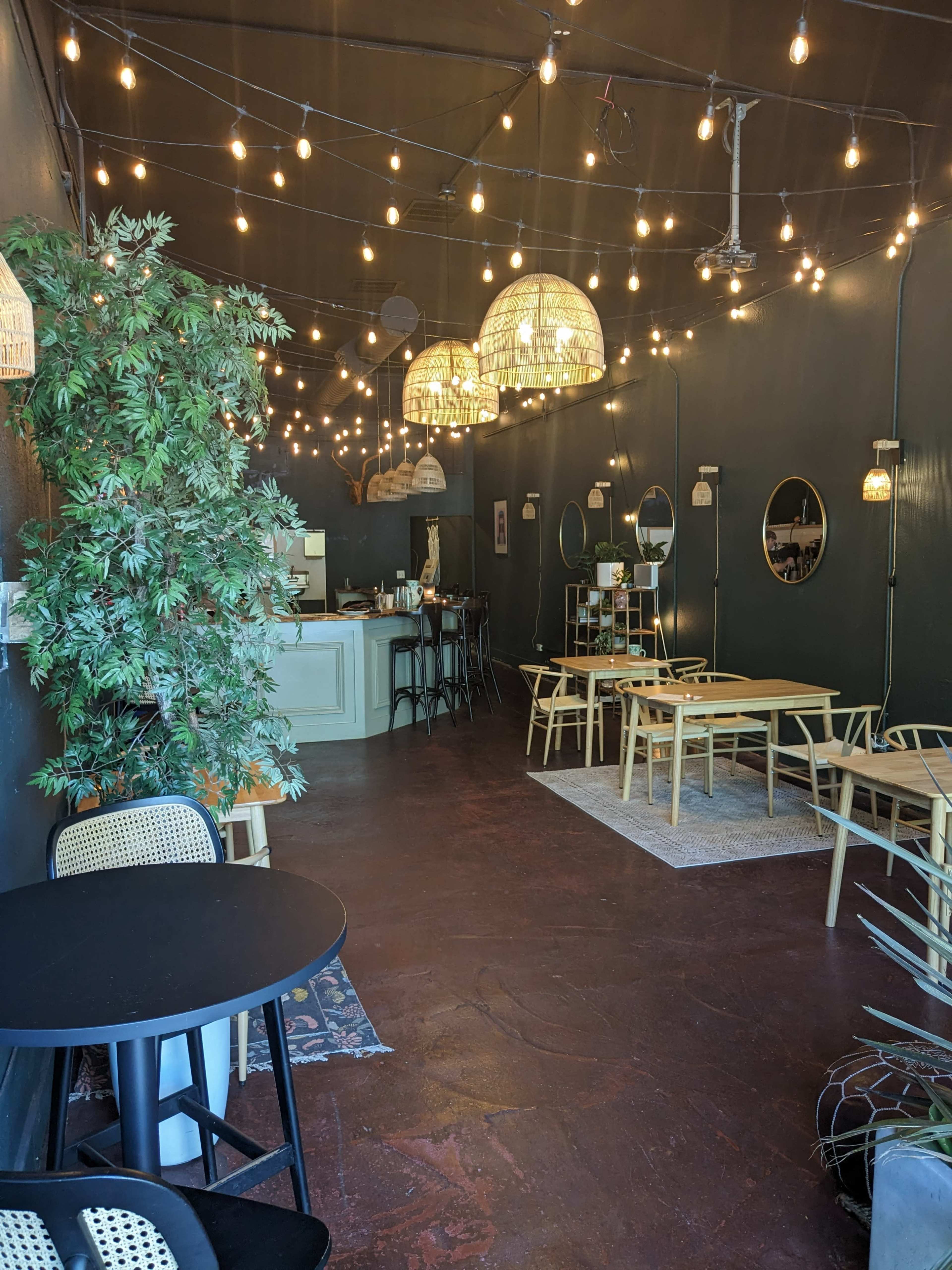 A cafe interior with string lights, wooden tables, chairs, and greenery, creating a cozy atmosphere.
