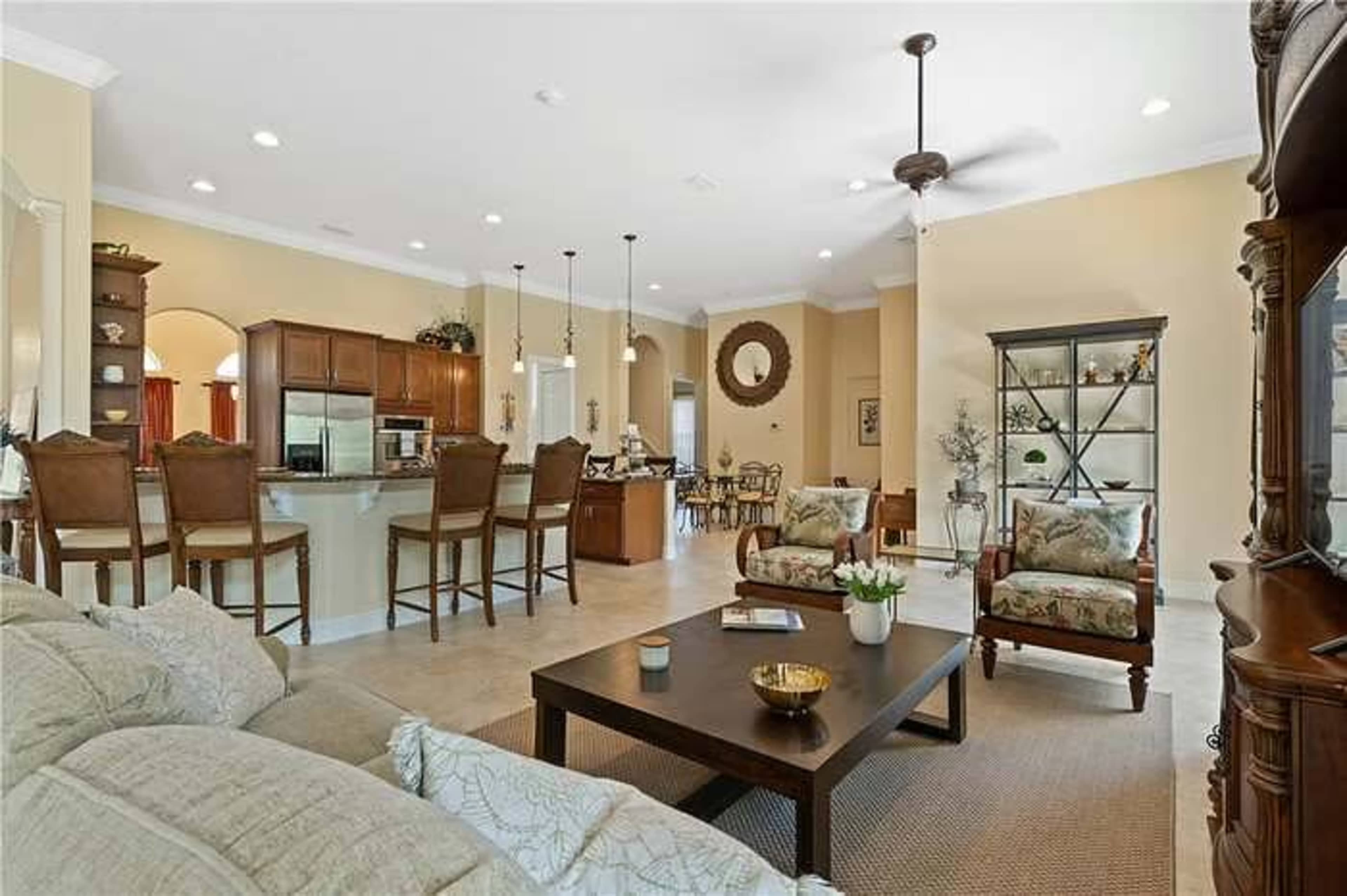 The image shows an open-concept living space with a kitchen, dining area, and seating arrangement, featuring light-colored walls and wood furnishings.