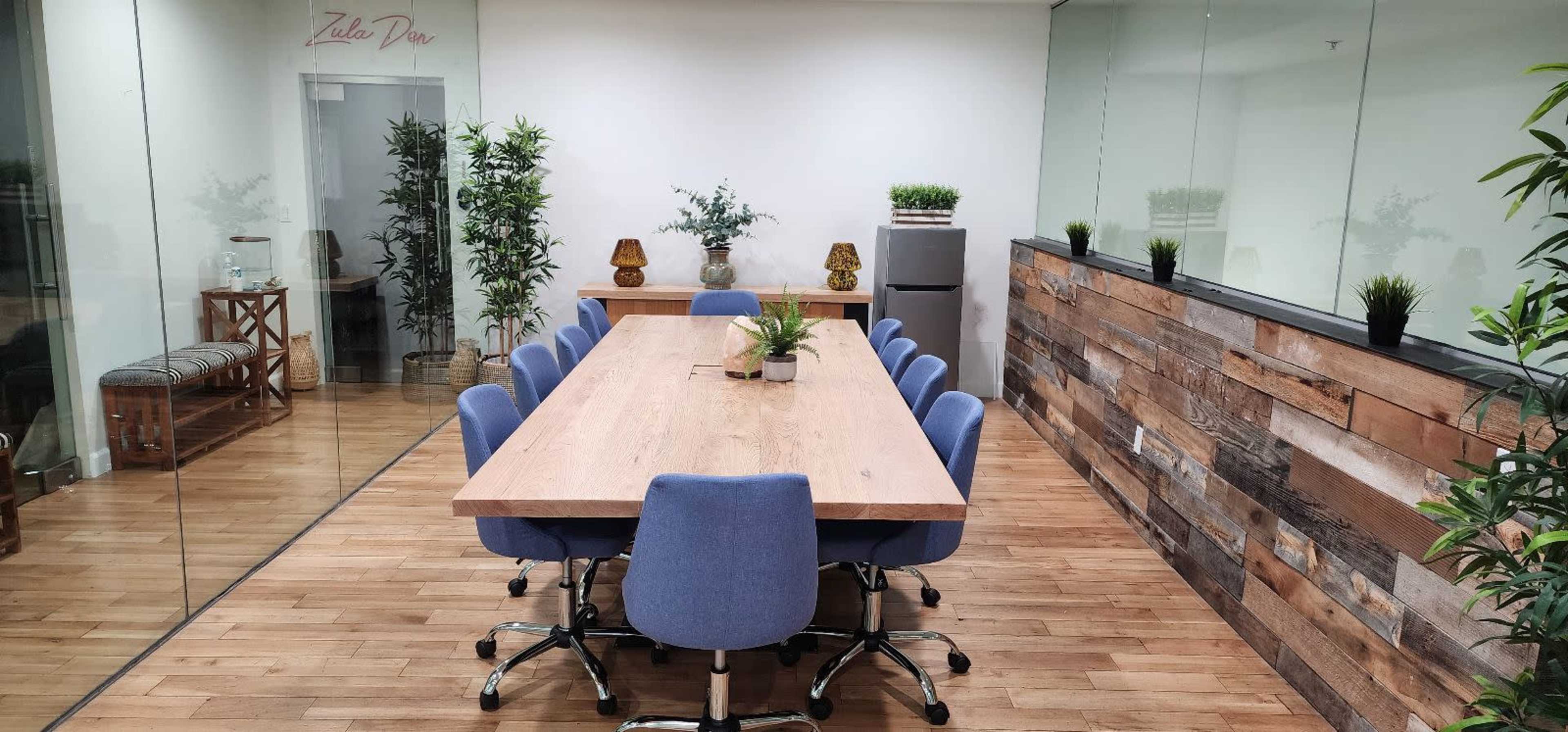 A modern conference room with a large wooden table surrounded by blue chairs, flanked by glass walls and decorative plants.