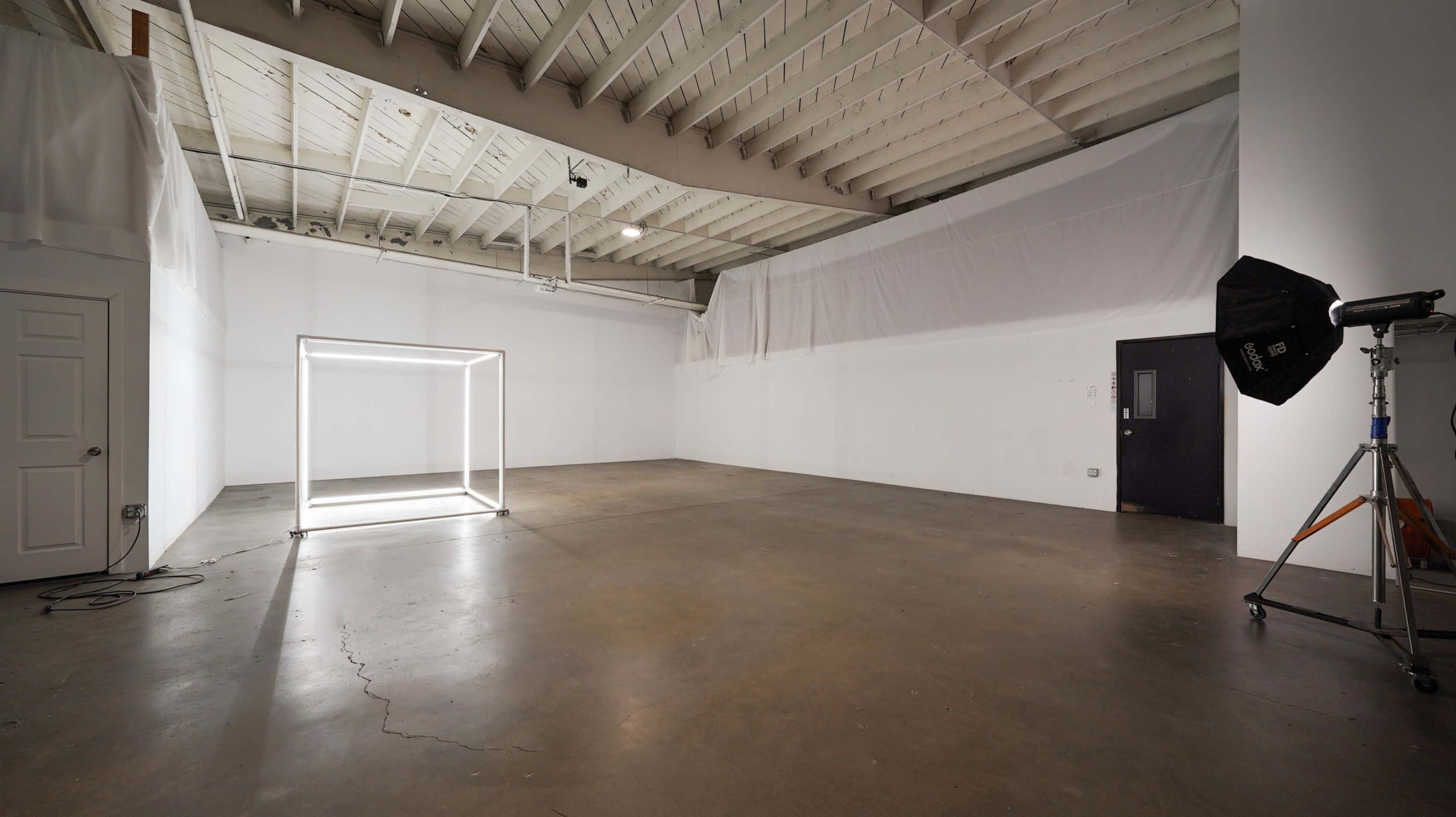 The image shows a spacious, empty gallery or studio with a large illuminated square frame in the center and white draping along the walls.
