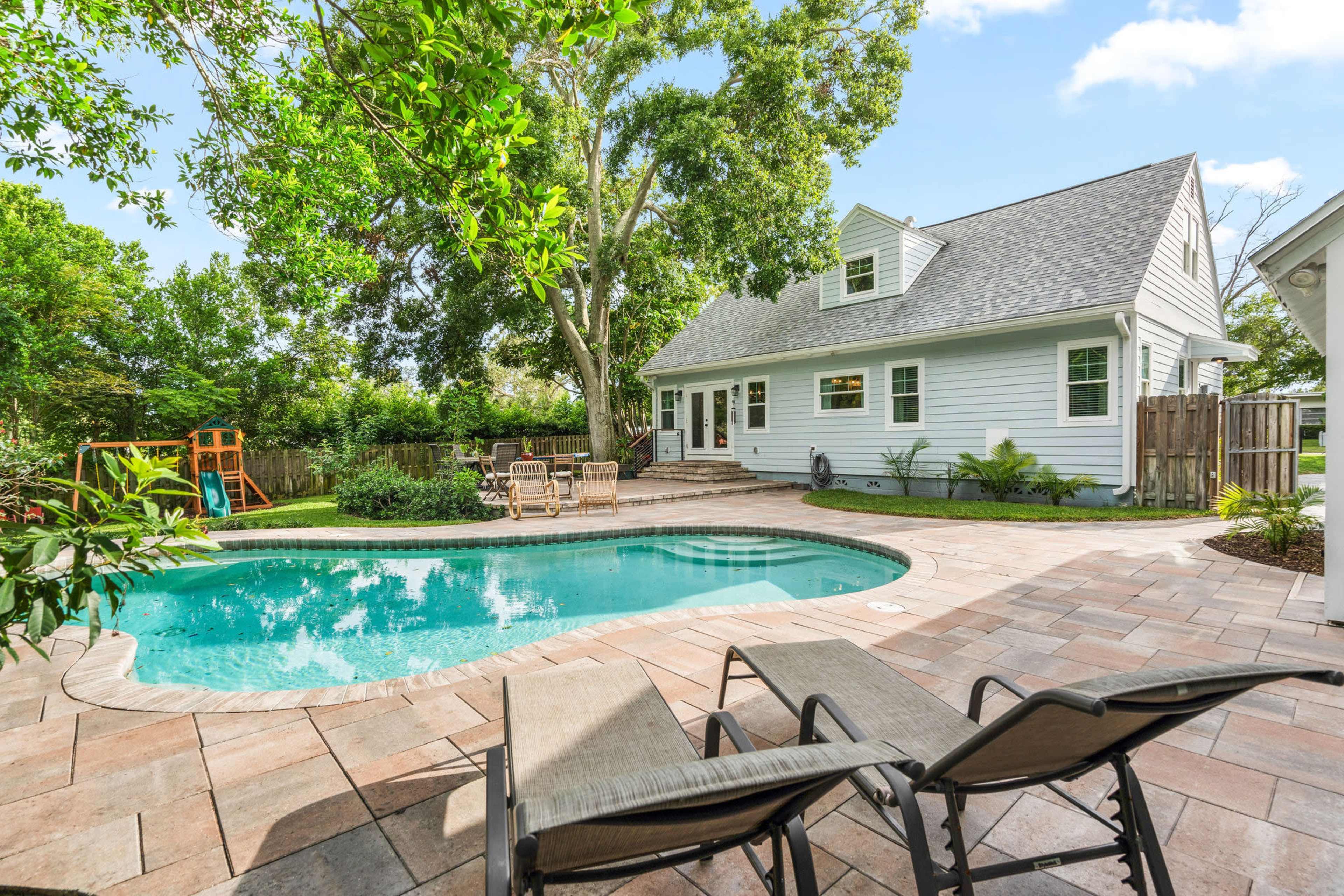 A backyard features a swimming pool surrounded by tiled decking, lounge chairs, and a playset, with a house visible in the background.
