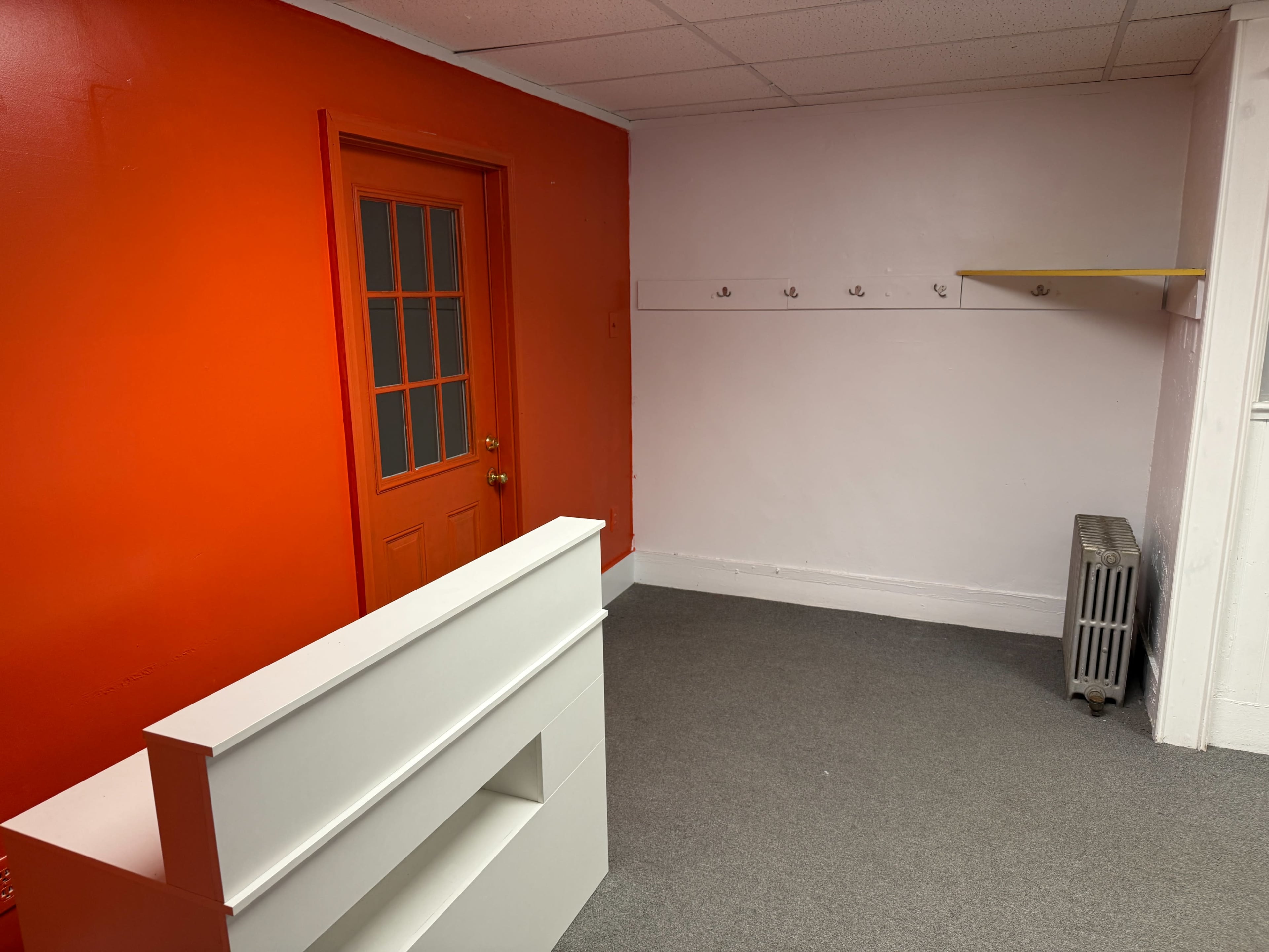 The image shows a sparsely furnished room with a vibrant red wall, a door, a coat rack, and a small shelf.