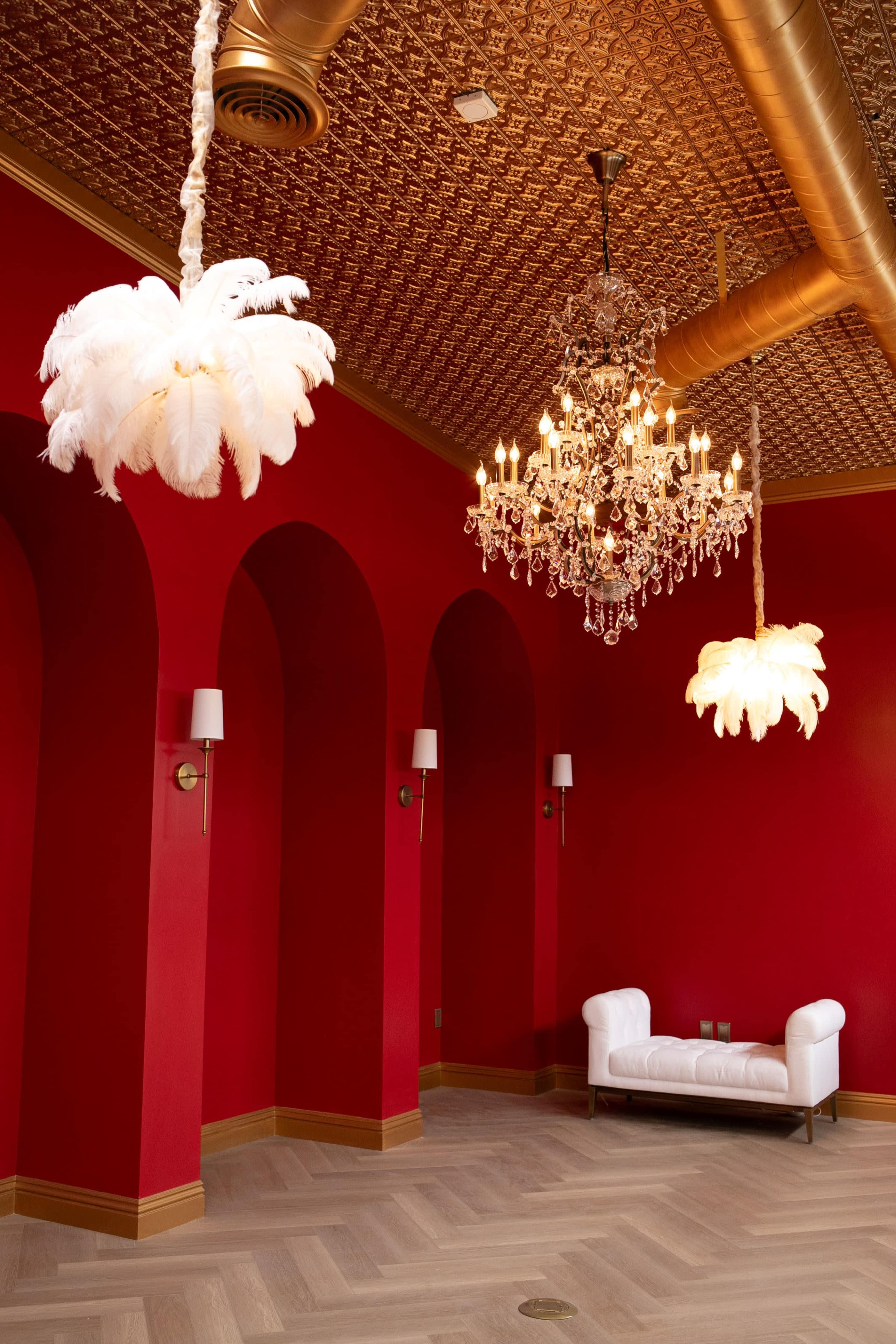 The room features a rich red wall with ornate golden ceiling, adorned with feather-like light fixtures and a crystal chandelier, along with a white sofa.