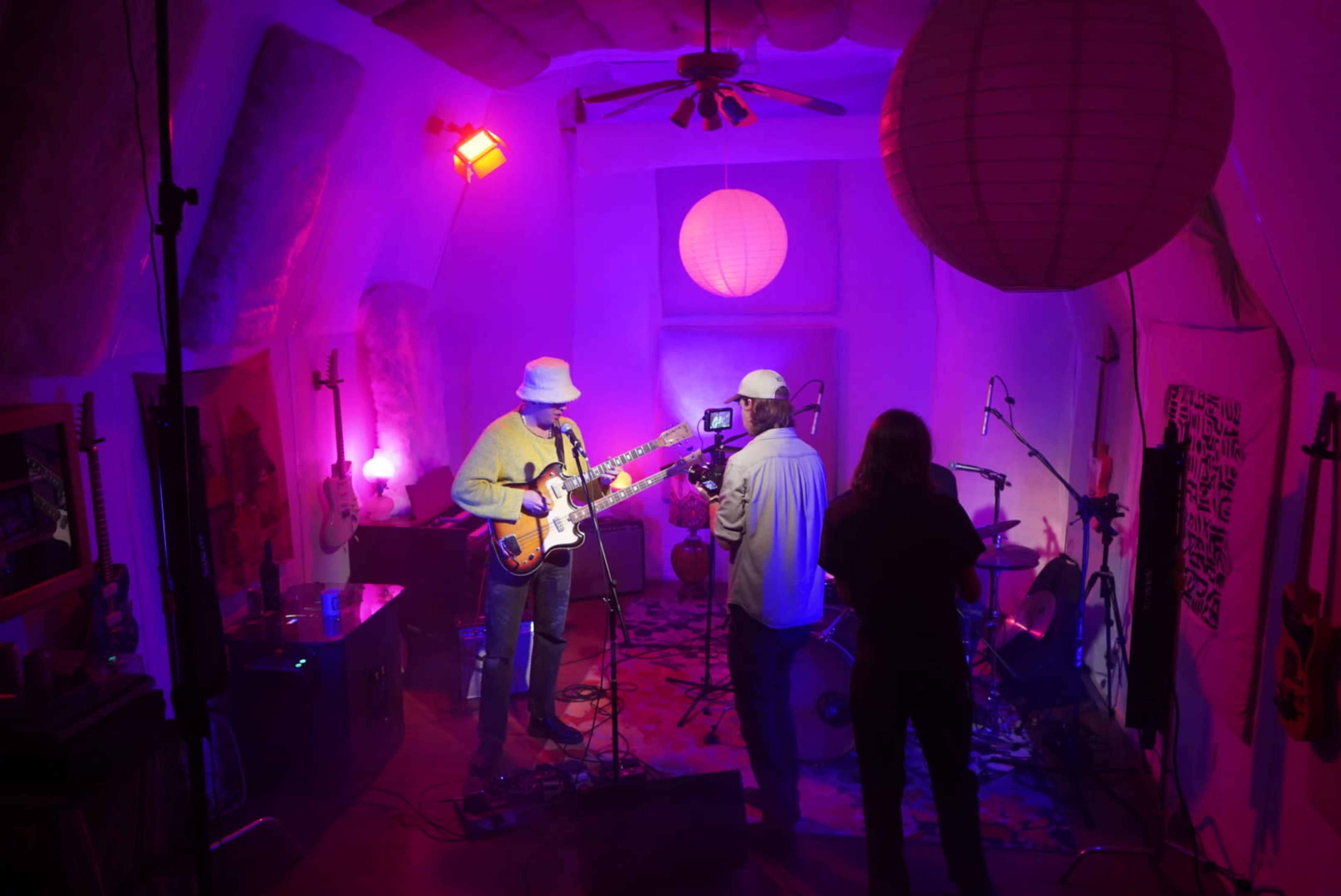 A musician performs on stage while being filmed in a colorful, dimly lit room adorned with paper lanterns and musical instruments.