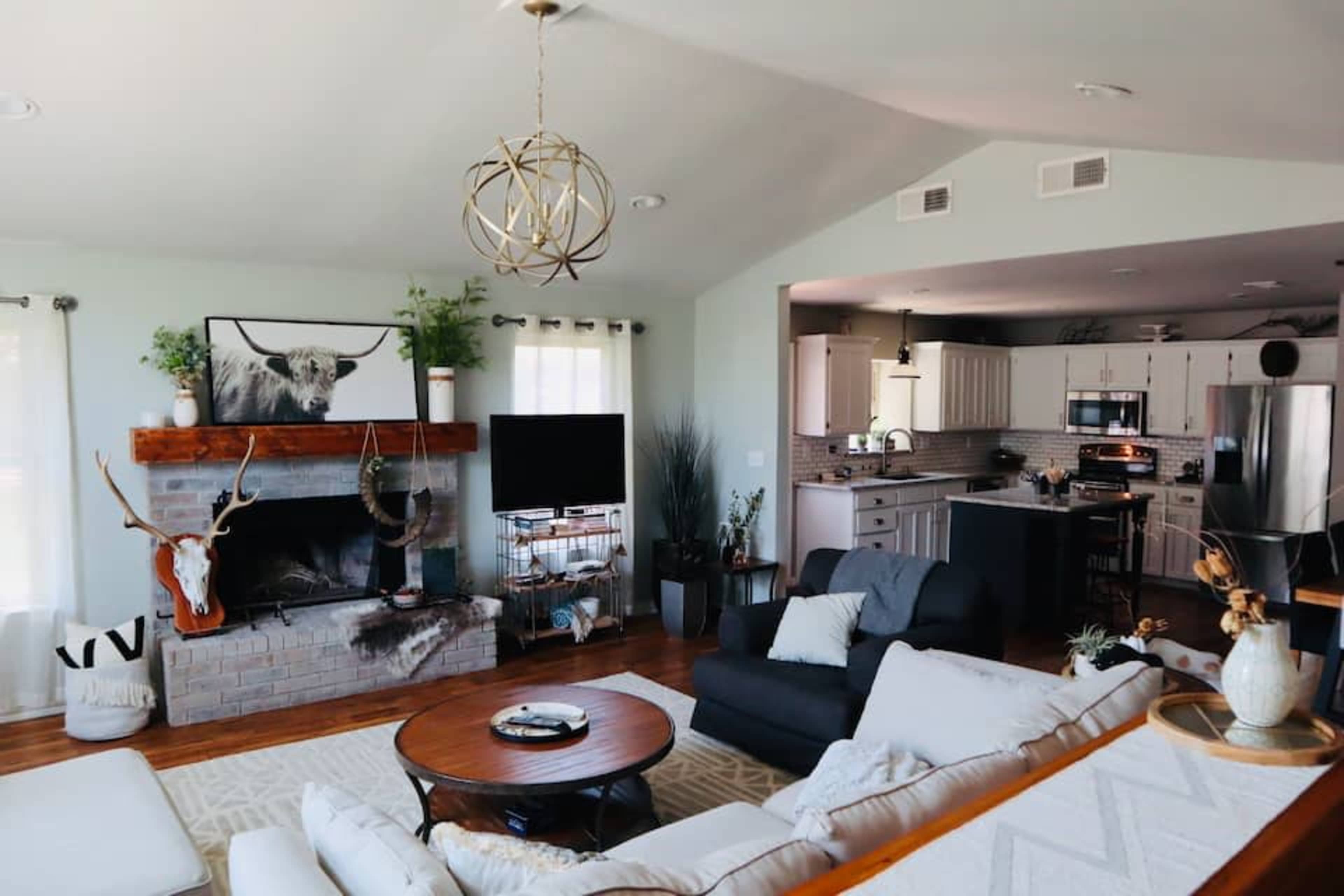 A spacious living room with a rustic fireplace, a television, and a kitchen area visible in the background.