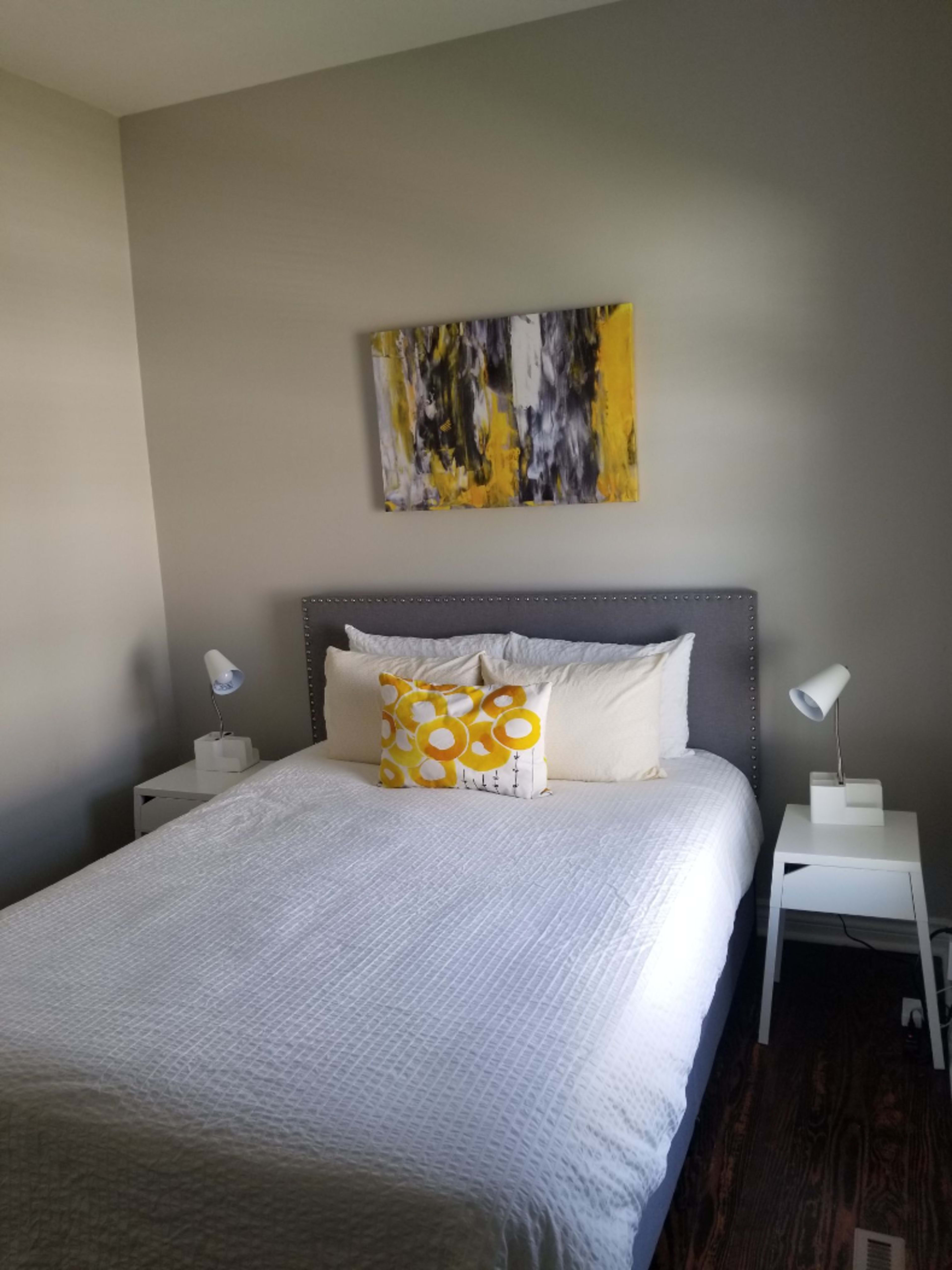 A neatly arranged bedroom featuring a gray upholstered bed with white bedding, yellow accent pillows, and two bedside tables, each with a lamp.