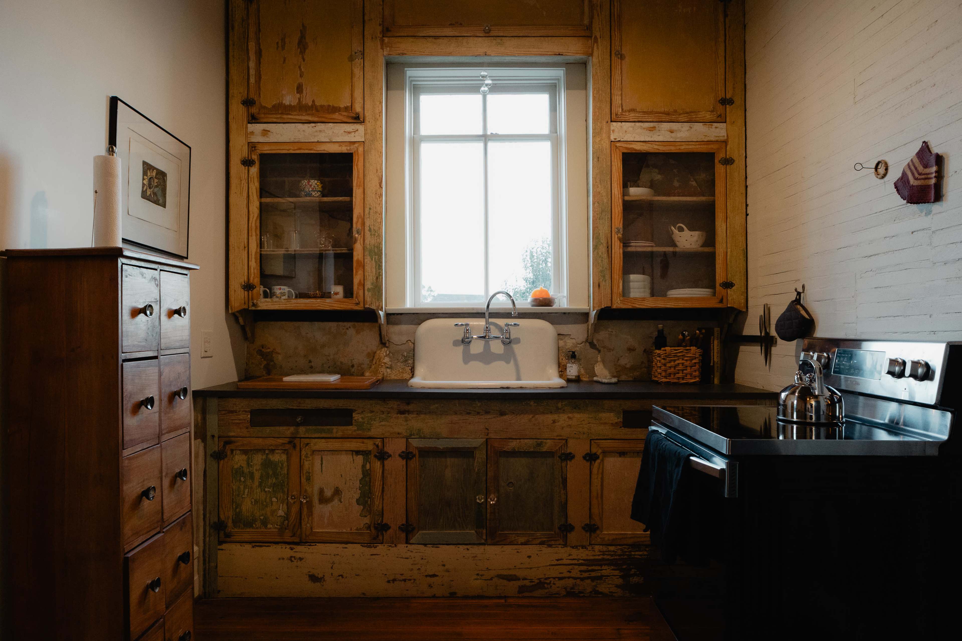 The image shows a rustic kitchen with weathered wooden cabinets, a large farmhouse sink, and a window above the sink allowing natural light to enter.