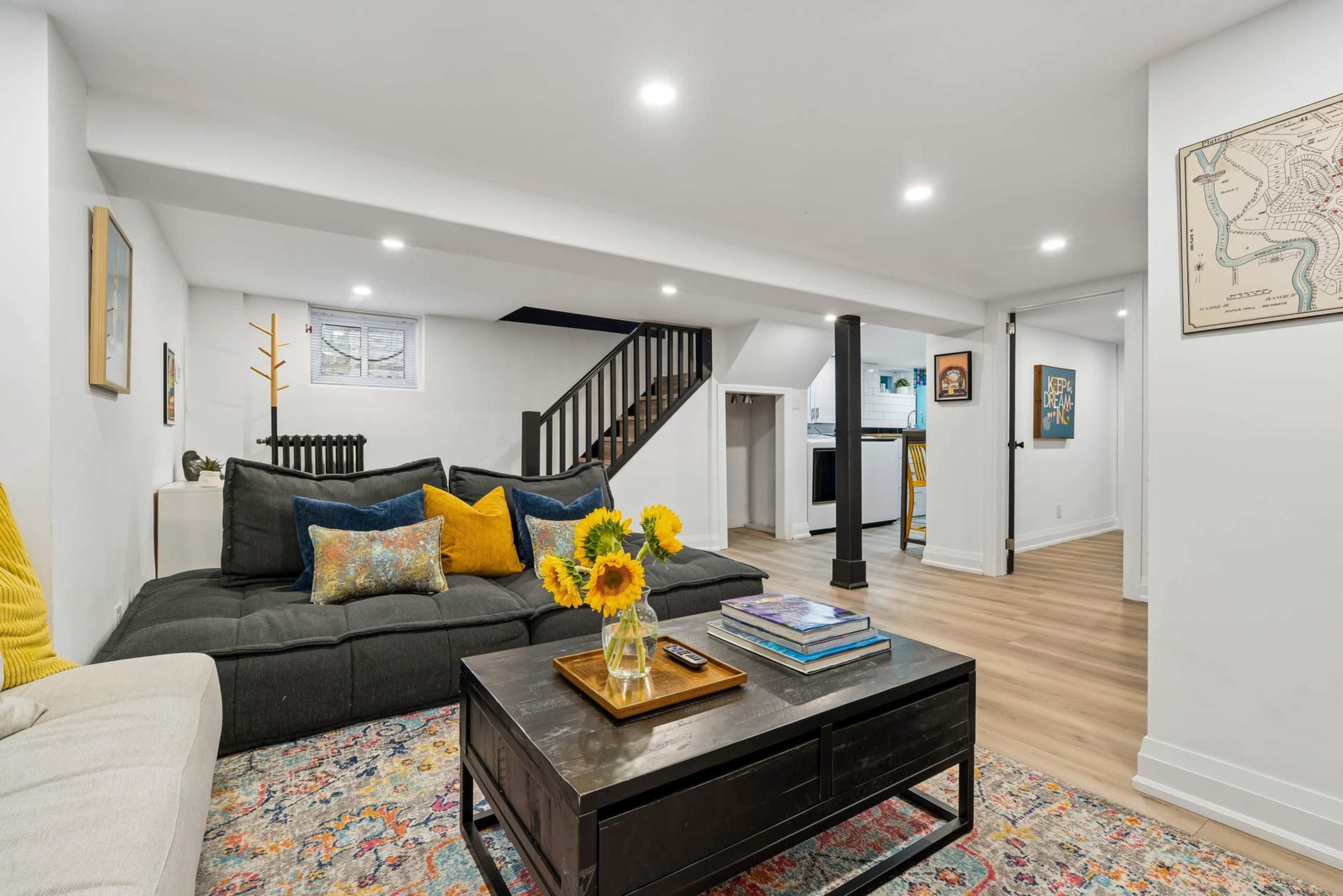 A cozy, well-lit basement living area features a dark sofa adorned with colorful cushions, a coffee table with a vase of sunflowers, and a staircase leading to an upper level.