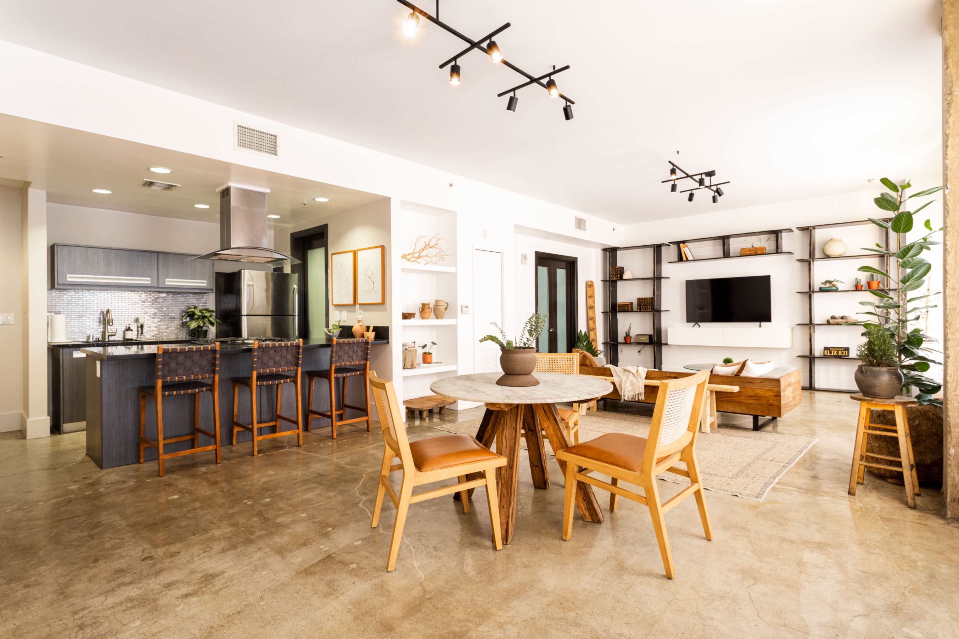 A modern open-concept living area featuring a kitchen with bar stools, a dining table, and a living space with shelves and a television.