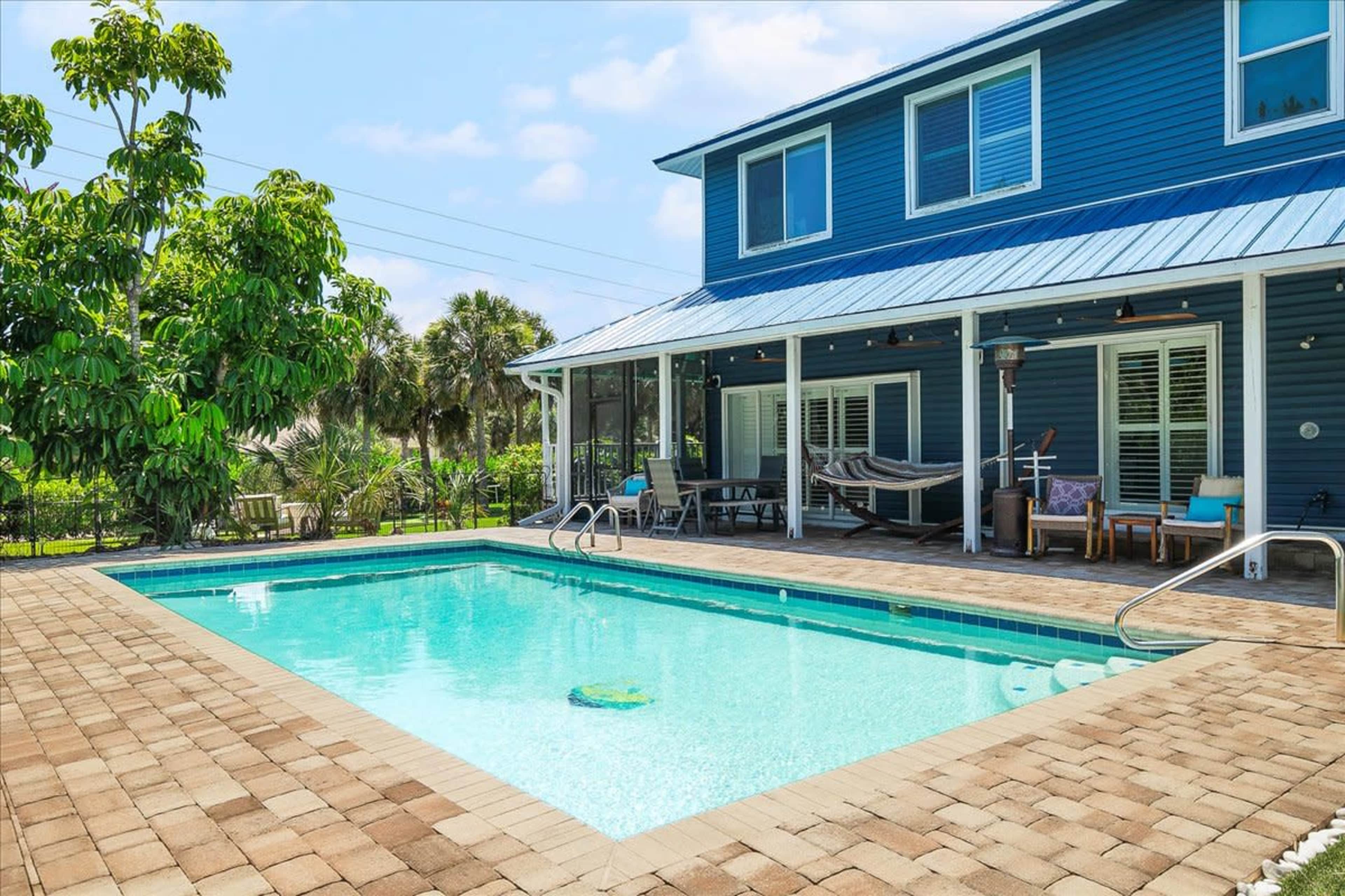 The image shows a blue two-story house with a screened porch overlooking a rectangular swimming pool surrounded by a paved patio.
