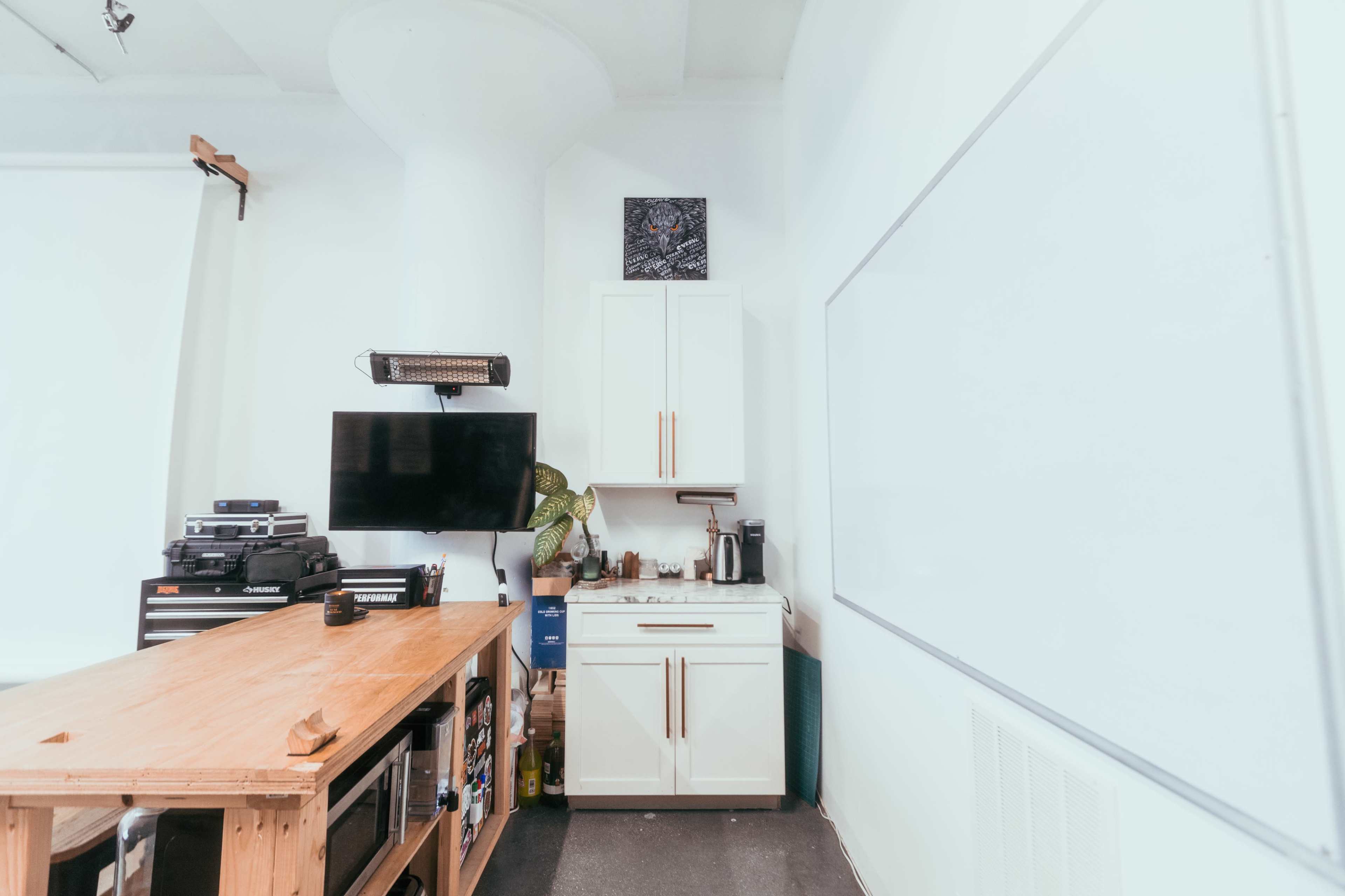 The image shows a well-lit room featuring a wooden table, a mounted television, cabinets, and a whiteboard against a white wall.