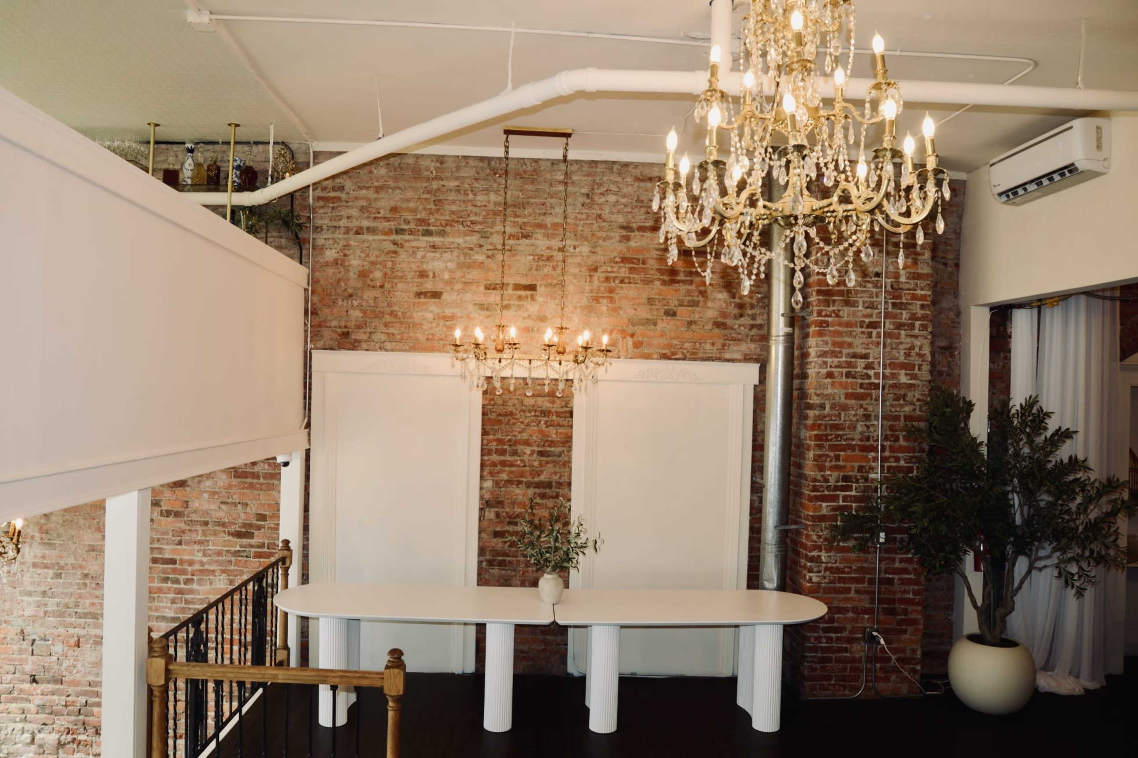 The scene features a modern interior with exposed brick walls, two chandeliers, and a white oval table decorated with a vase of greenery.