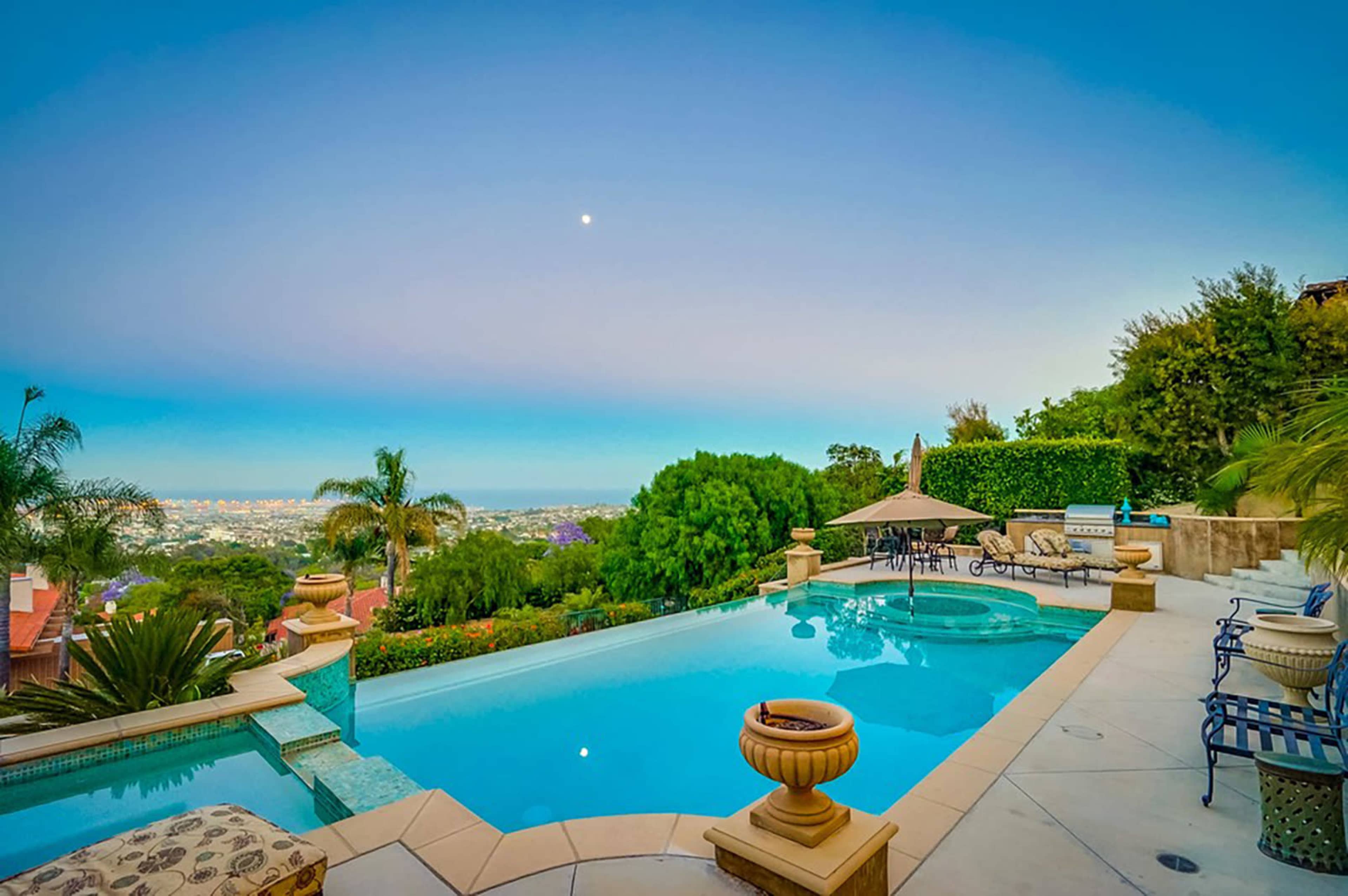 A swimming pool with stone edges and decorative planters, surrounded by lush greenery and a view of the city skyline under a clear sky.