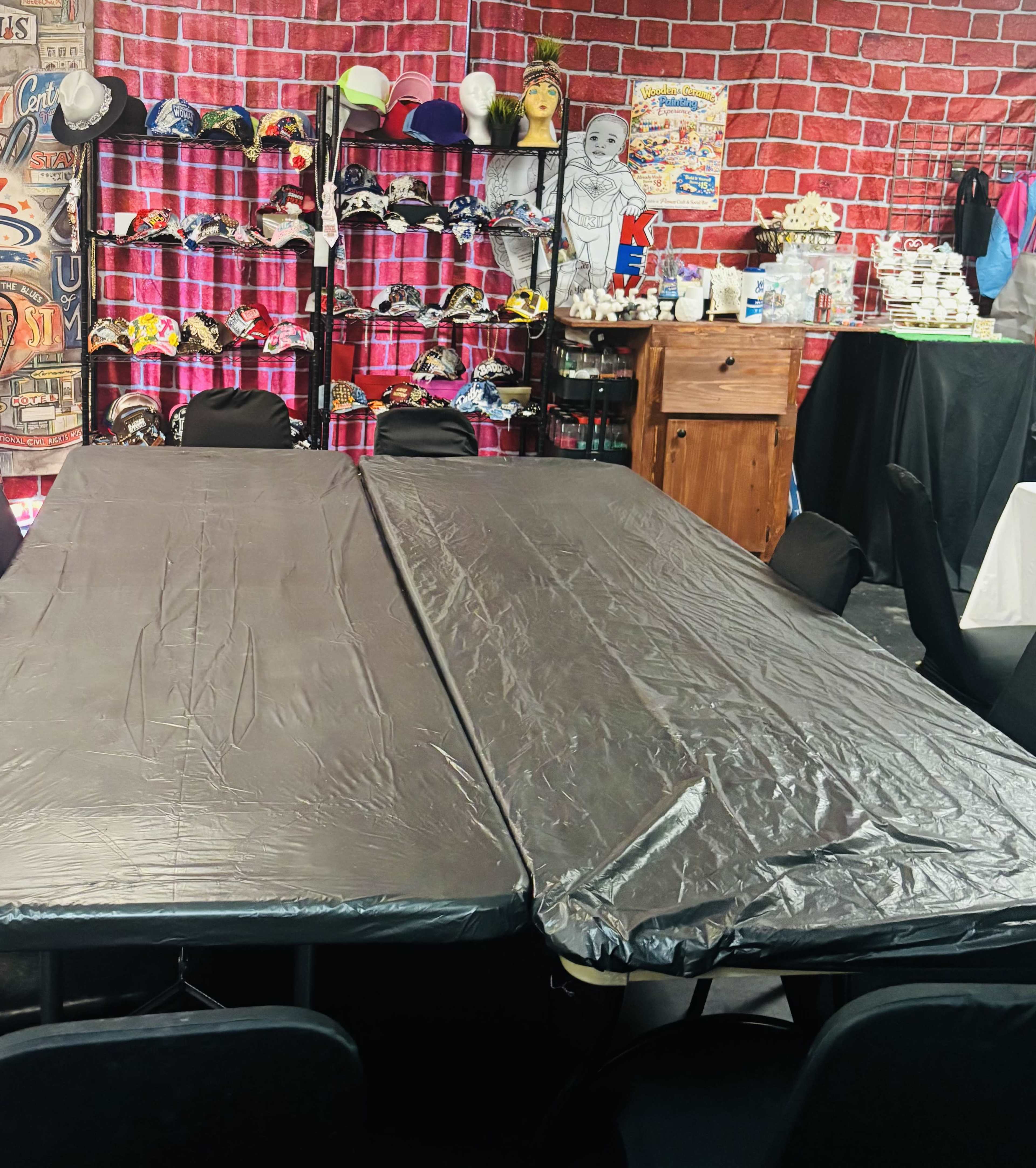 The image shows a set of two long tables covered with black plastic tablecloths, surrounded by chairs, with a backdrop displaying shelves filled with colorful hats and other items.