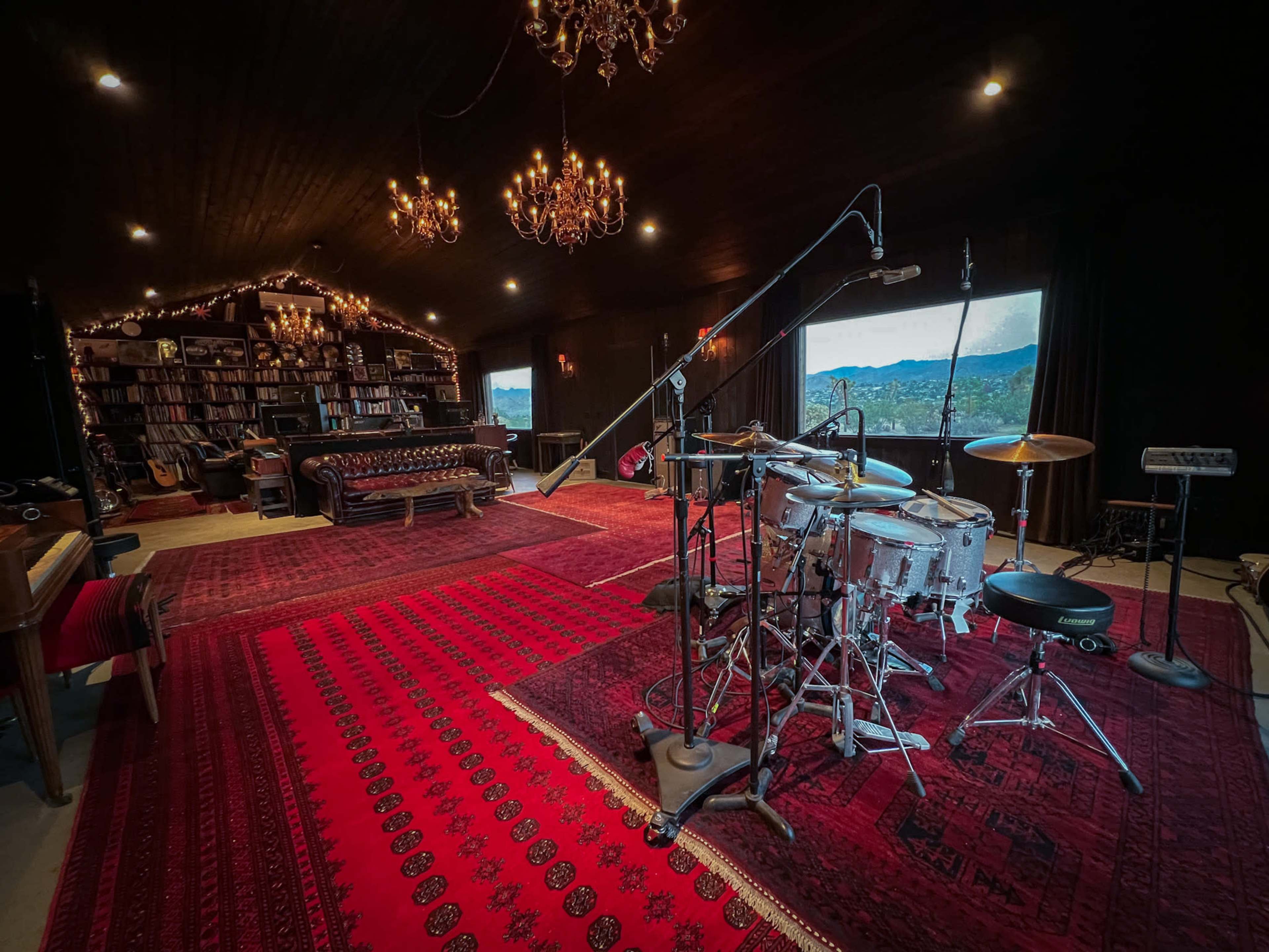 The image shows a music studio with a drum set in the foreground, surrounded by dark wood walls, chandeliers, and shelves filled with books.
