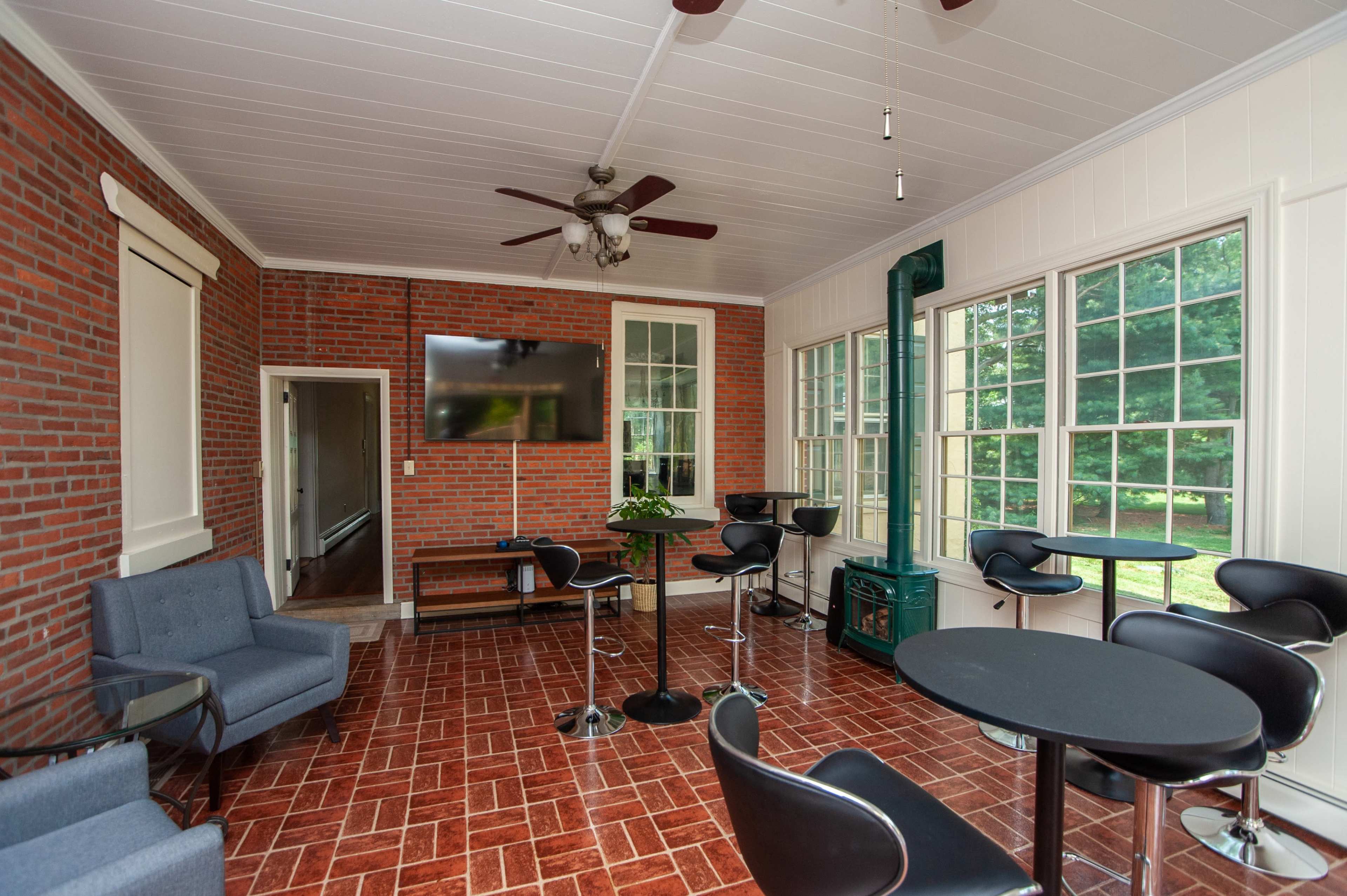 The image shows a sunroom with brick walls, large windows, and a mix of seating arrangements, including bar stools and a small table.