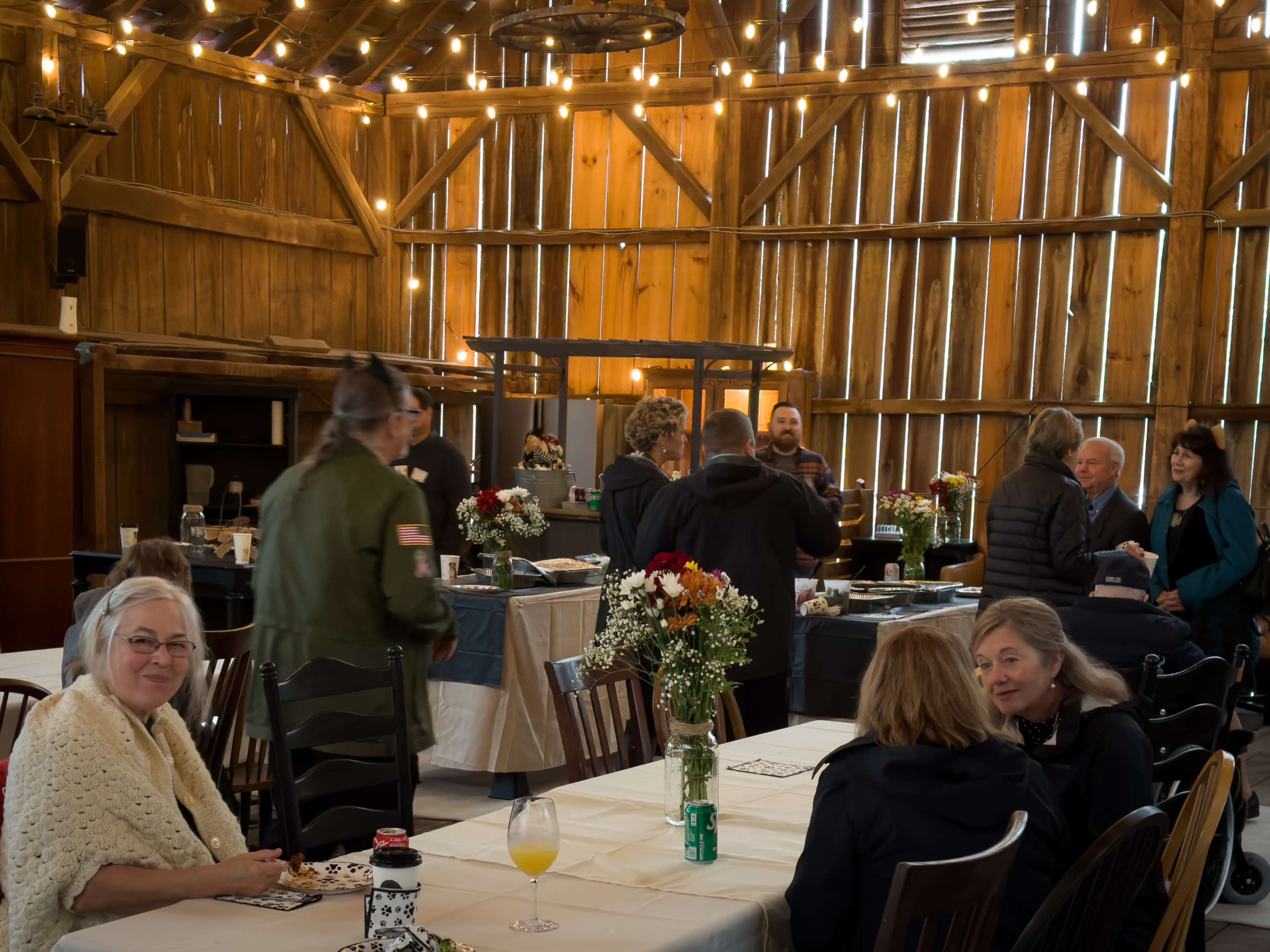 A group of people mingles and converses in a rustic barn setting decorated with string lights and wooden beams.