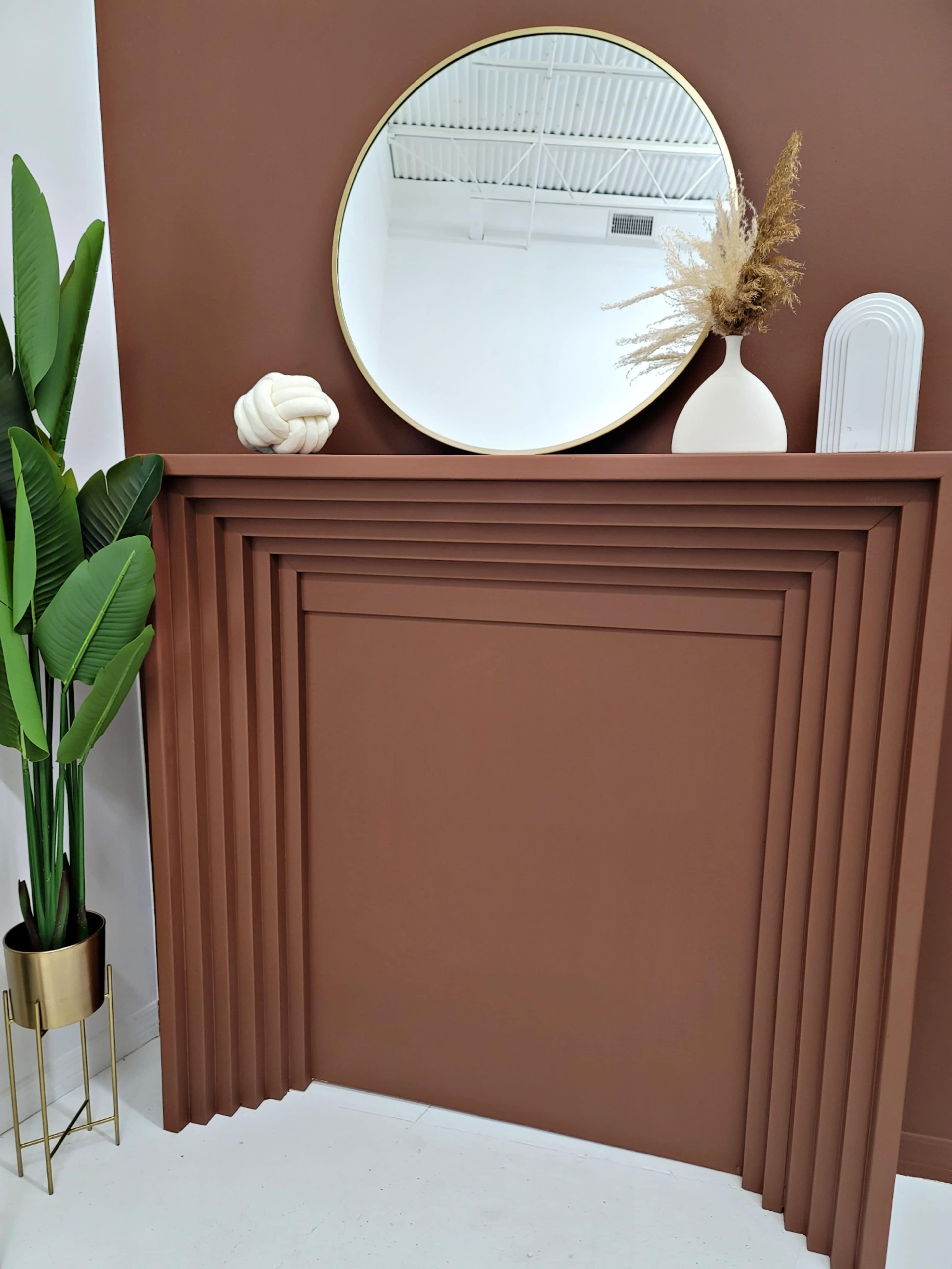 The image features a modern wall arrangement with a round mirror above a contoured brown mantel, complemented by a tall plant and decorative objects.