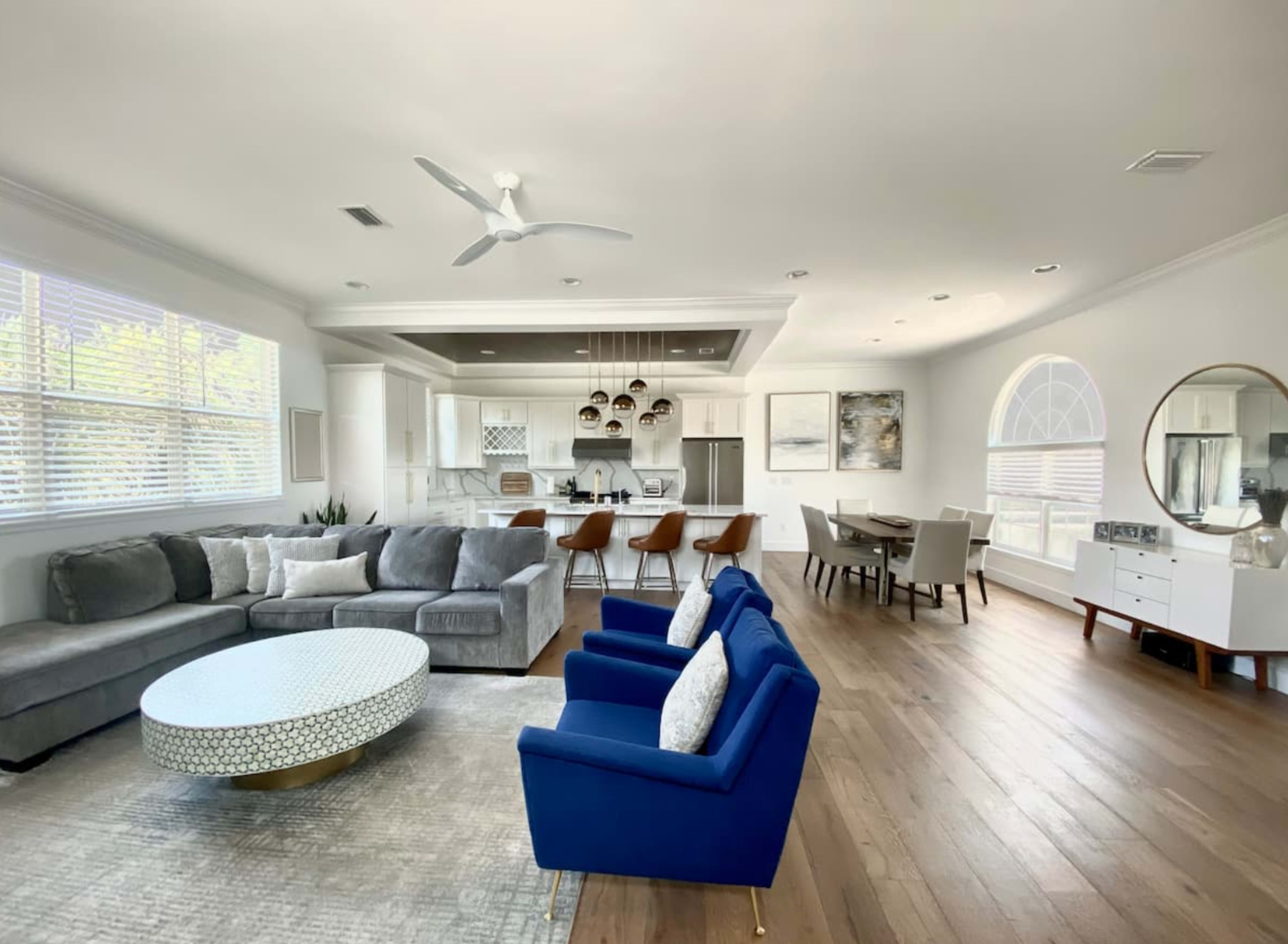 A modern living room features a sectional sofa, blue accent chairs, a circular coffee table, and an open layout connected to a dining area and kitchen.