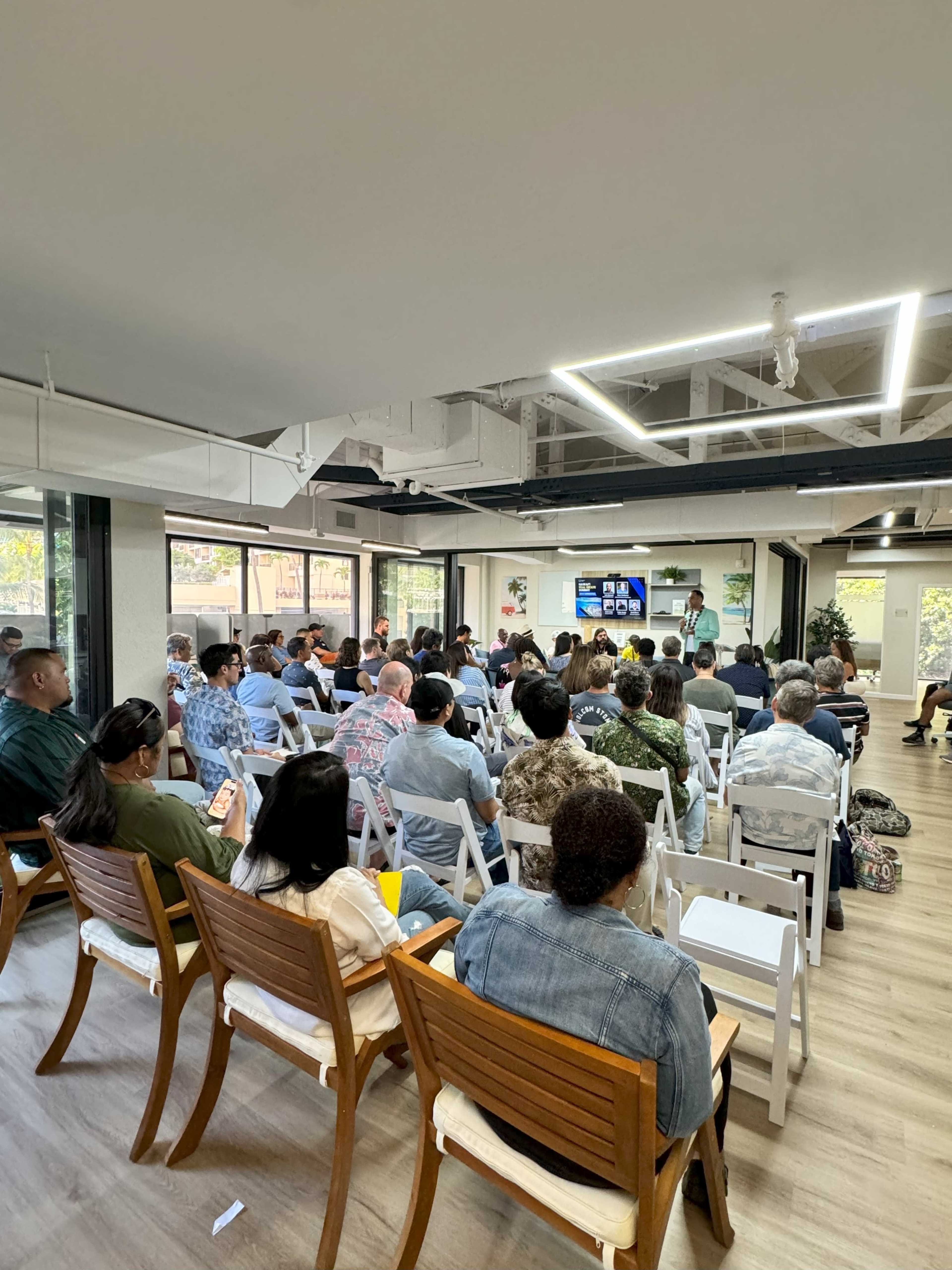 A group of attendees sits in rows of chairs facing a presenter and a screen in a modern meeting space.
