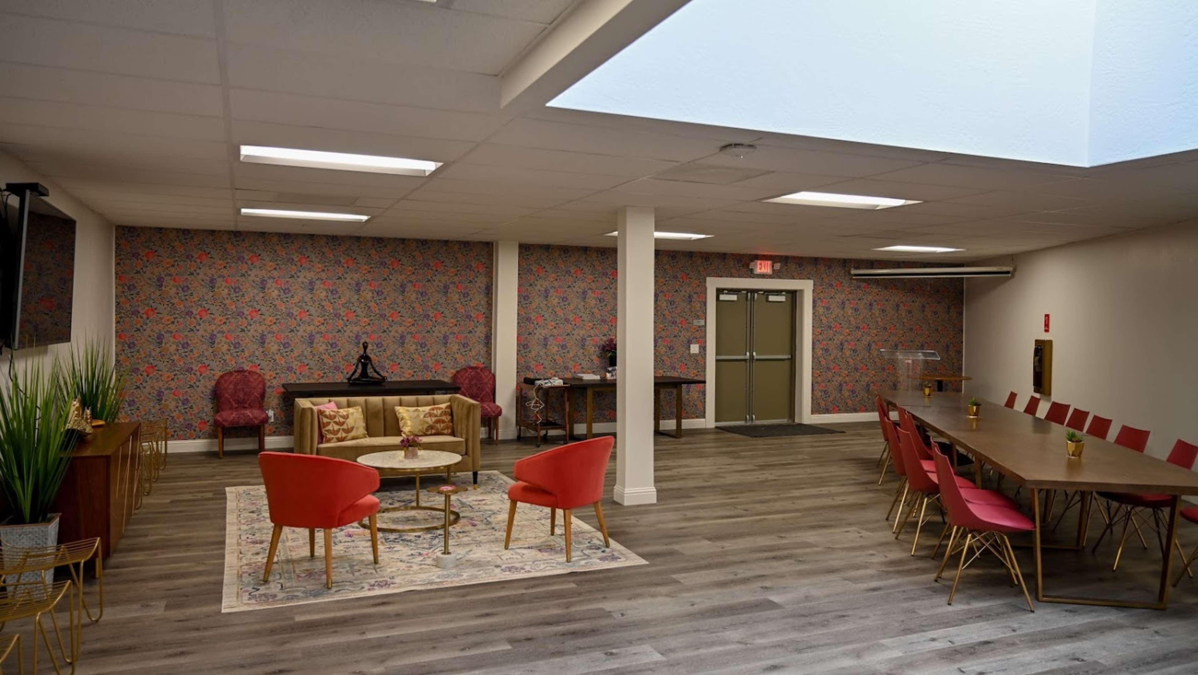 The interior of a spacious room features a mixture of seating arrangements, including red chairs and a long dining table, with decorative wallpaper and natural lighting from a skylight.