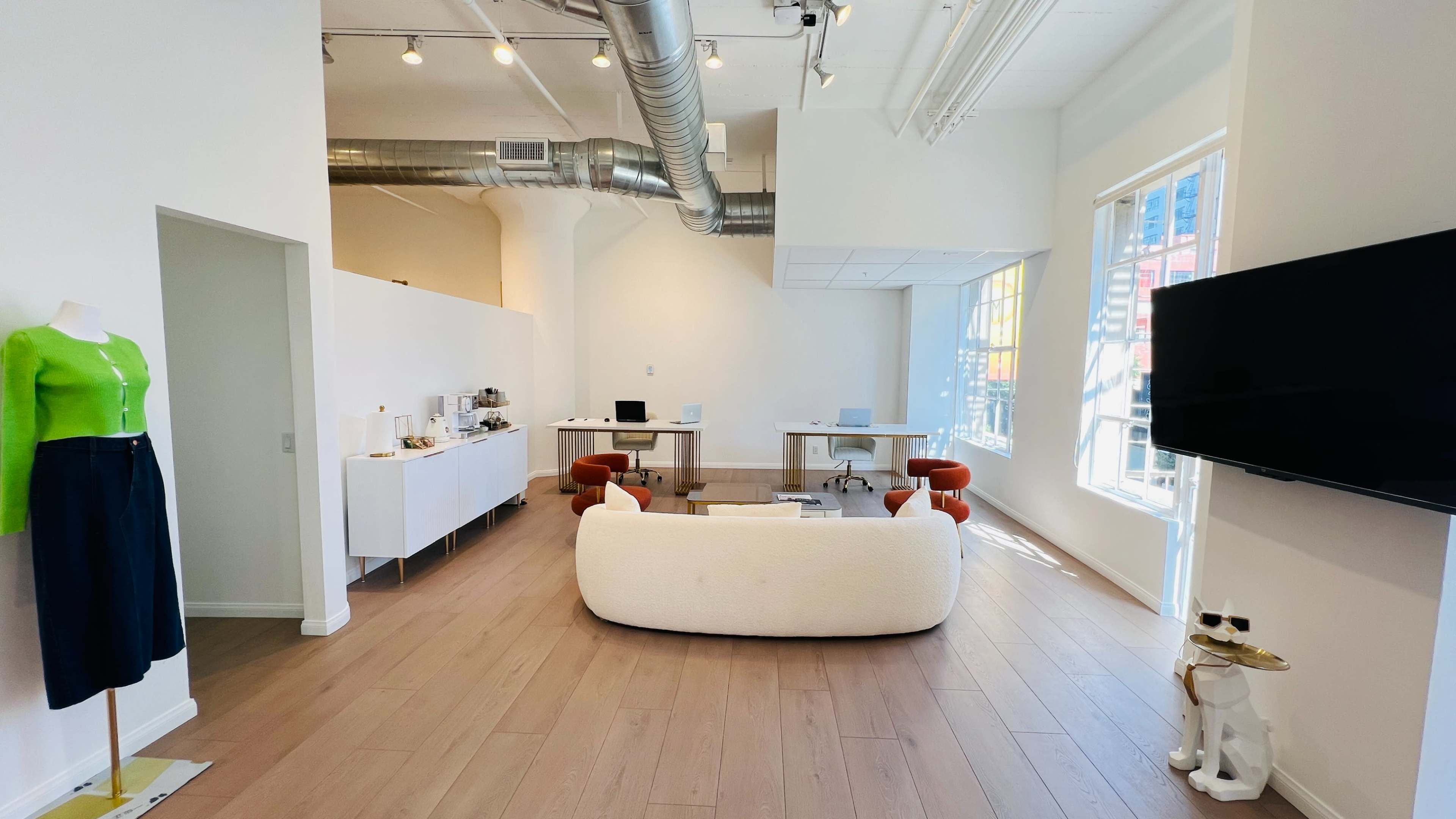 The image shows a modern office space featuring a white sofa, a mannequin dressed in green and dark blue, and desks set up with computers against white walls and large windows.